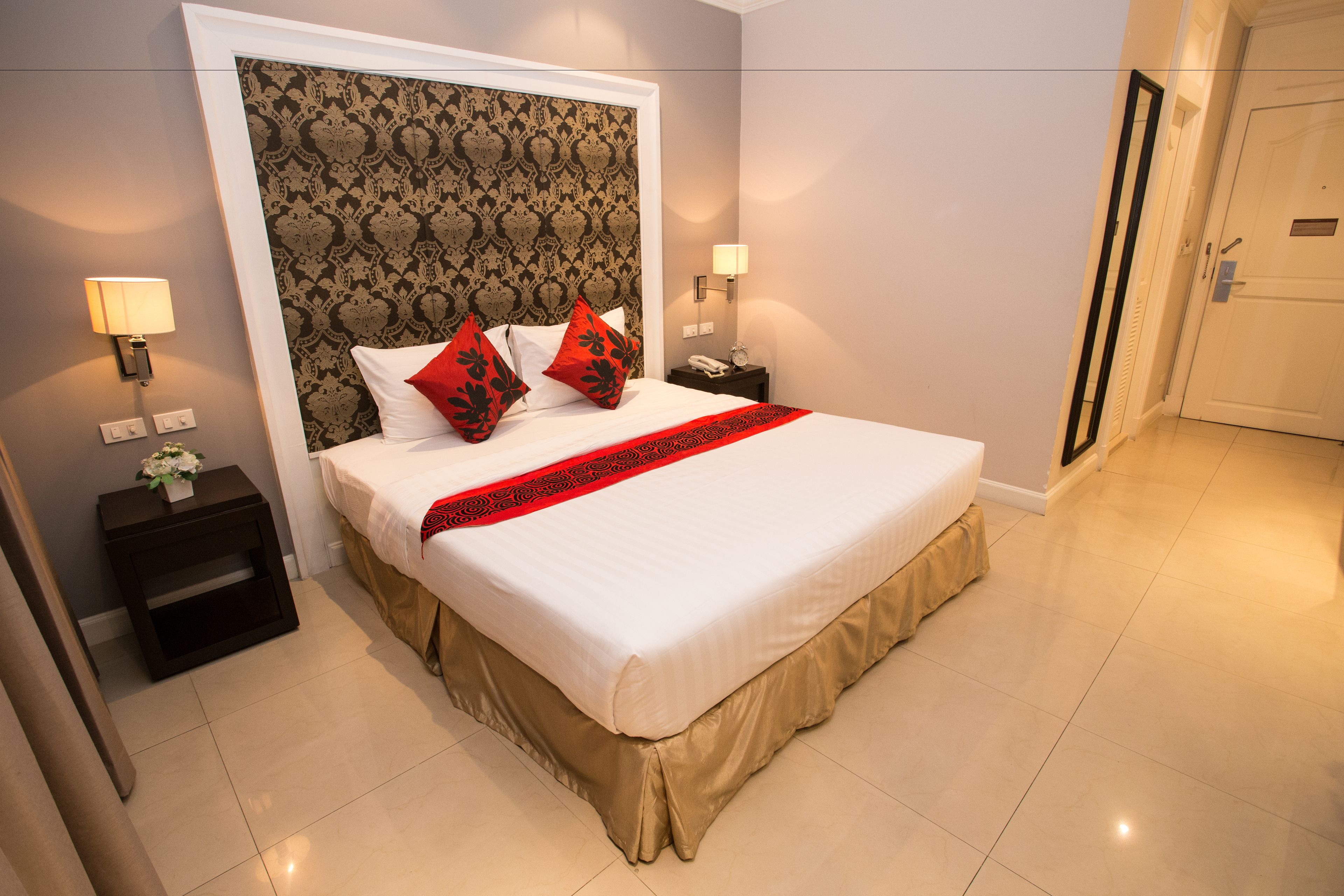 Una cama bien hecha con ropa blanca y detalles rojos, un cabecero oscuro con un diseño estampado, y una mesita de noche con una lámpara y un florero.