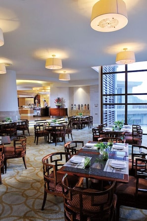 A spacious hotel dining area with multiple tables and chairs, large windows offering a view of the city, and a visible kitchen area in the background.