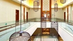 Un pasillo amplio de hotel con barandilla de vidrio, múltiples lámparas colgantes de estilo jaula y una puerta de madera central.