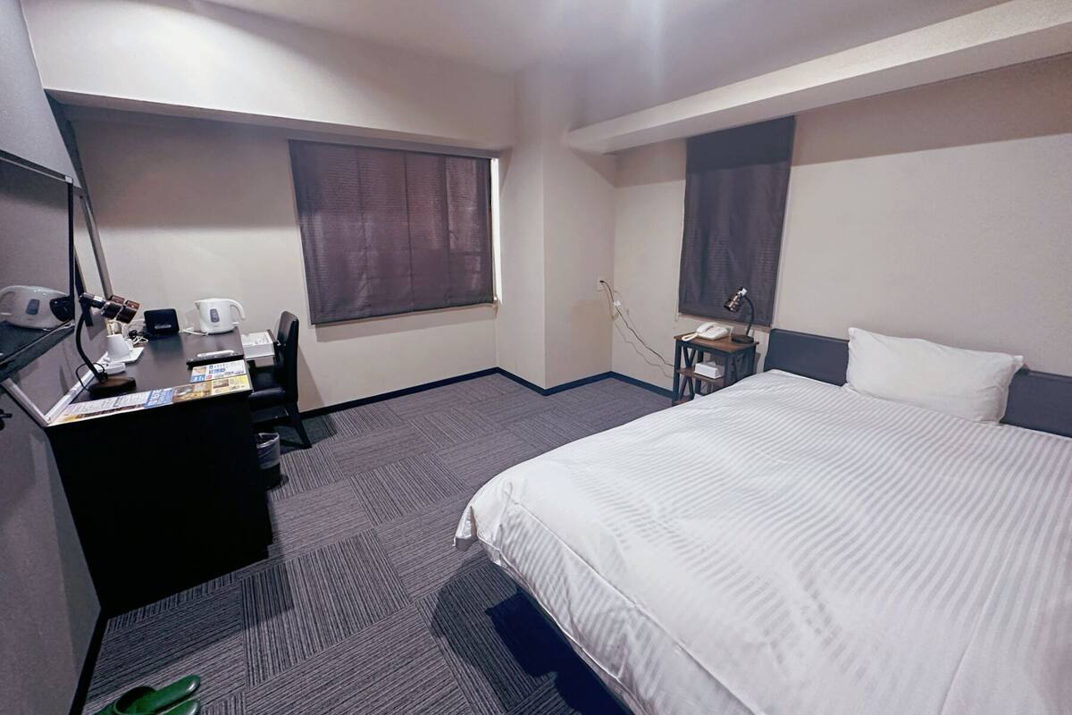 A hotel room with a bed, a desk with a computer and phone, a chair, and a window with curtains.