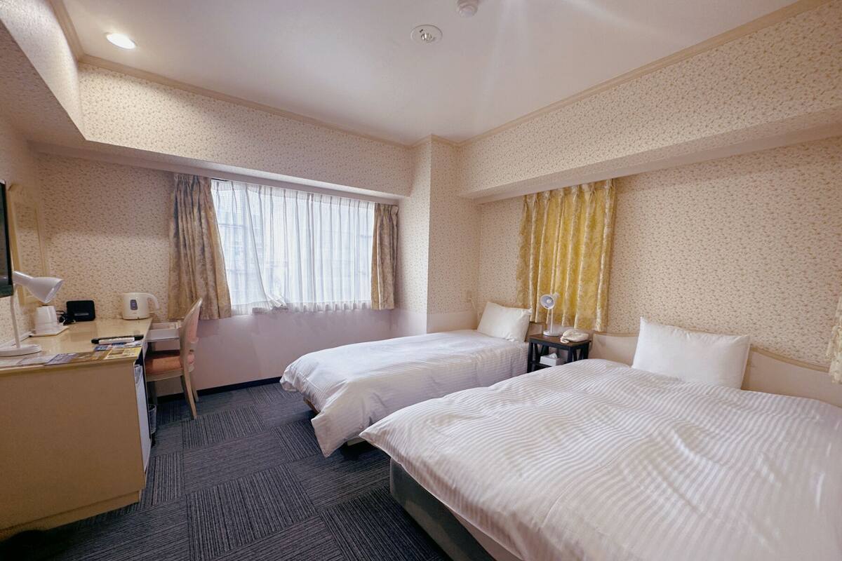 A hotel room with two beds, a desk with a chair, a desk lamp, a telephone, and a window with curtains.