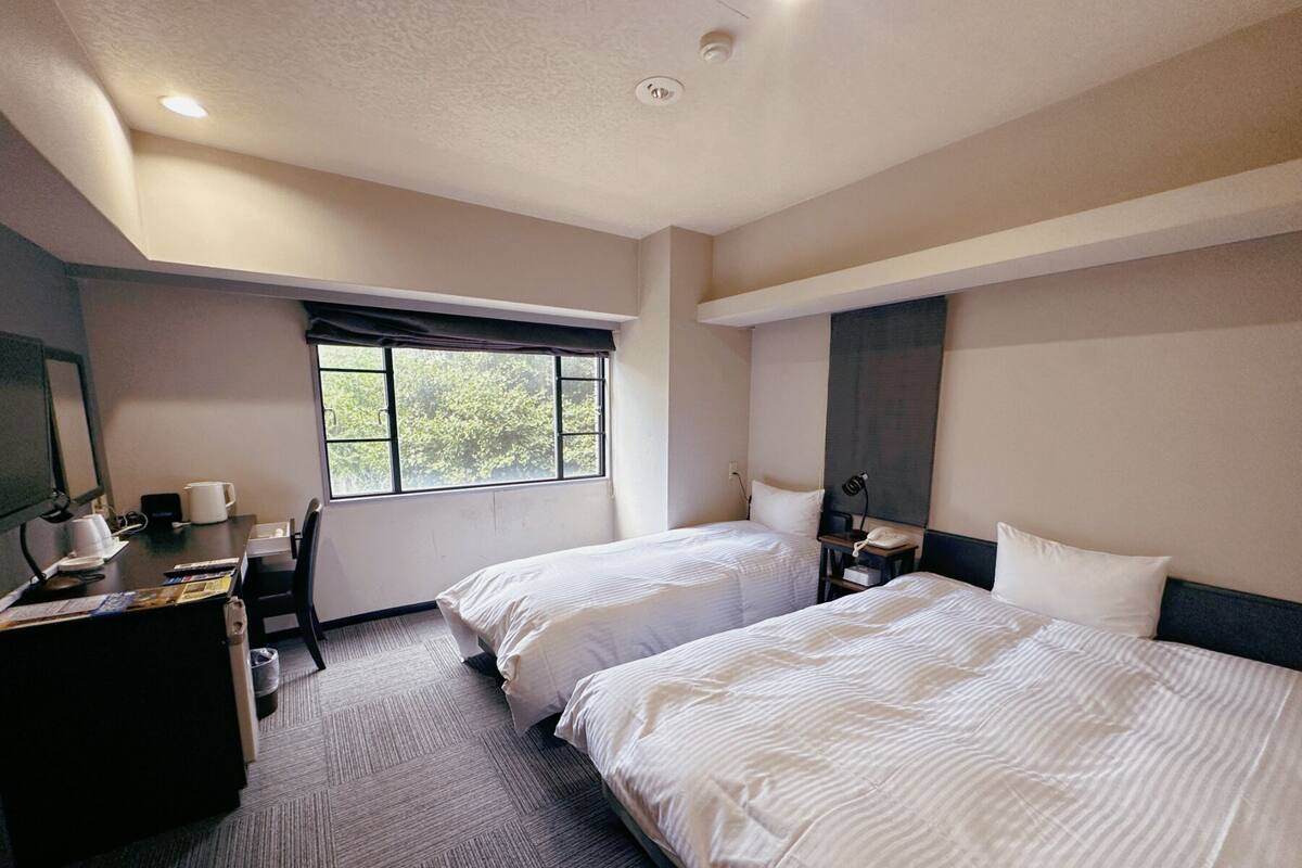 A hotel room with two beds, a desk, a chair, a window with a view of trees, and a ceiling light.