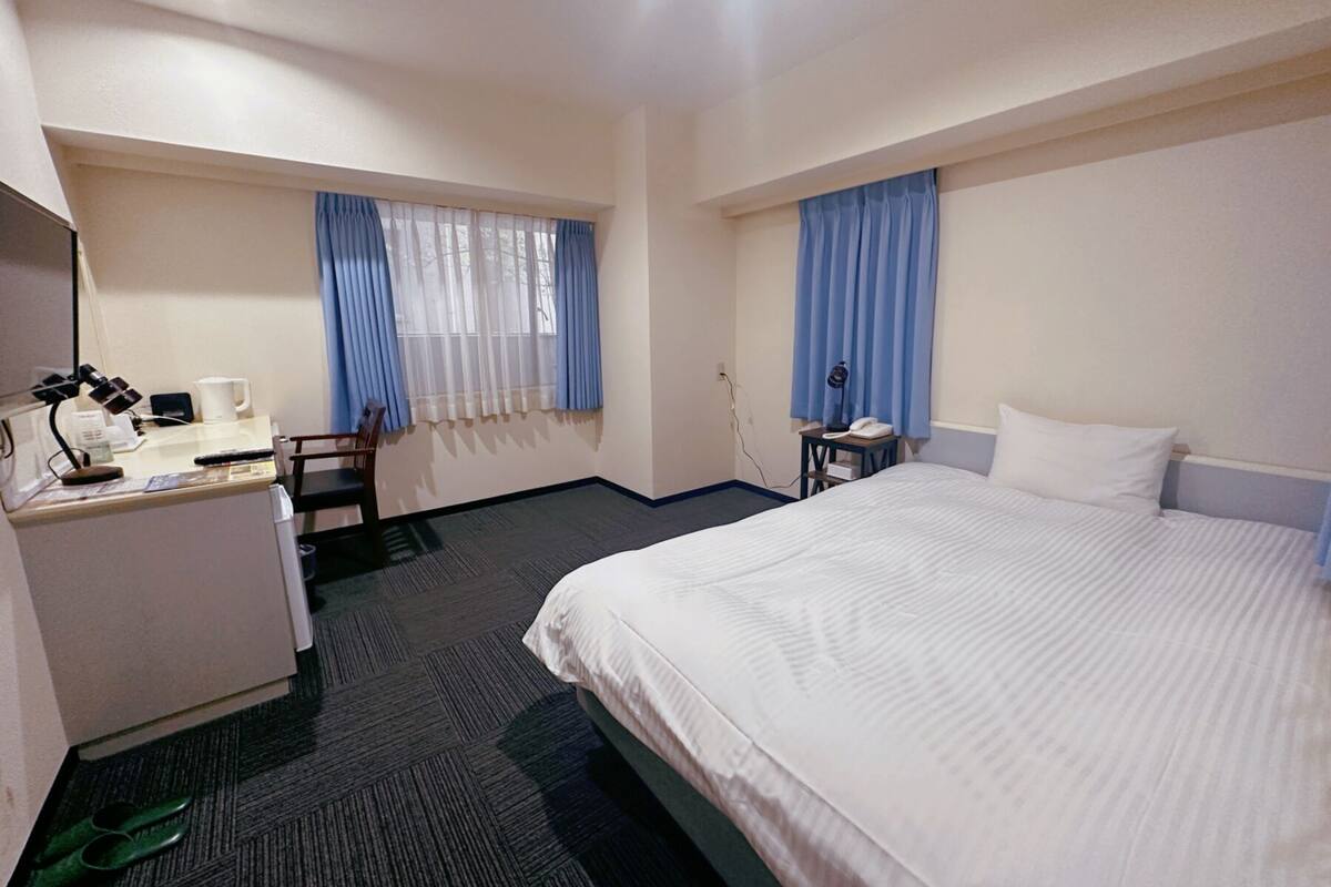 A hotel room with a bed, desk, chair, and a small table with a phone and lamp.