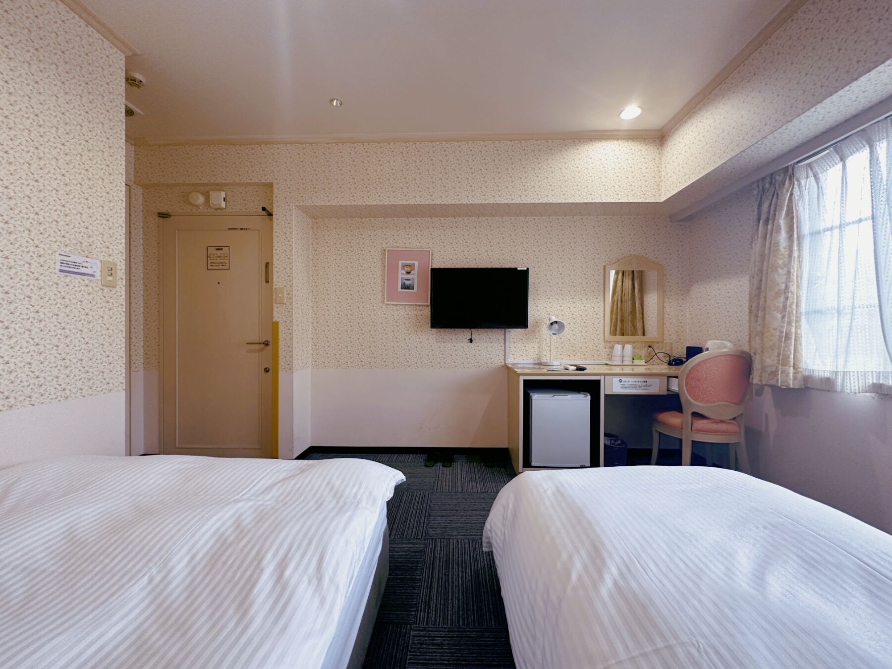 A hotel room with two beds, a desk with a computer, a chair, a TV, and a window with curtains.