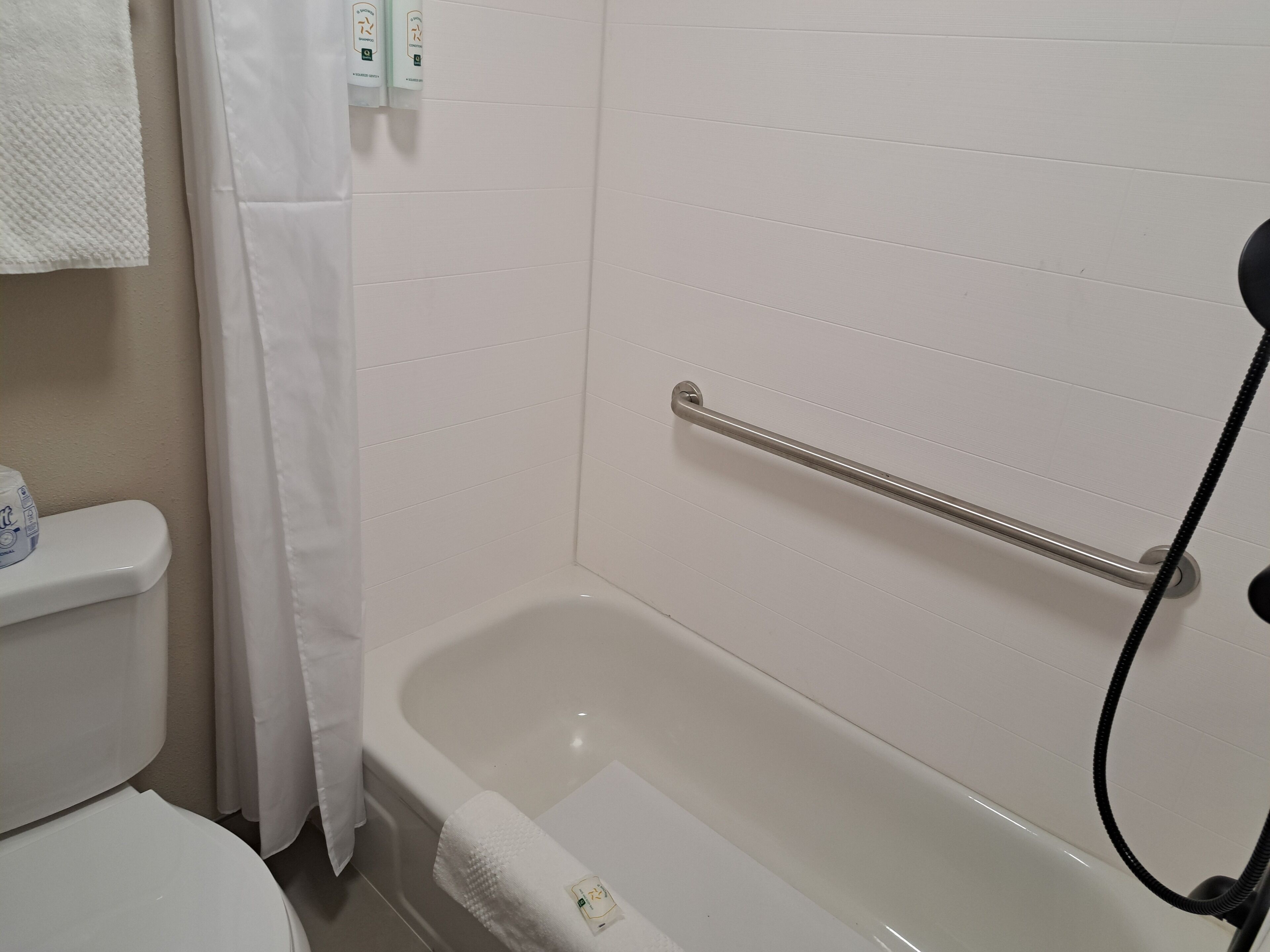 A bathroom with a white bathtub, a handrail, a toilet, and a shower curtain.