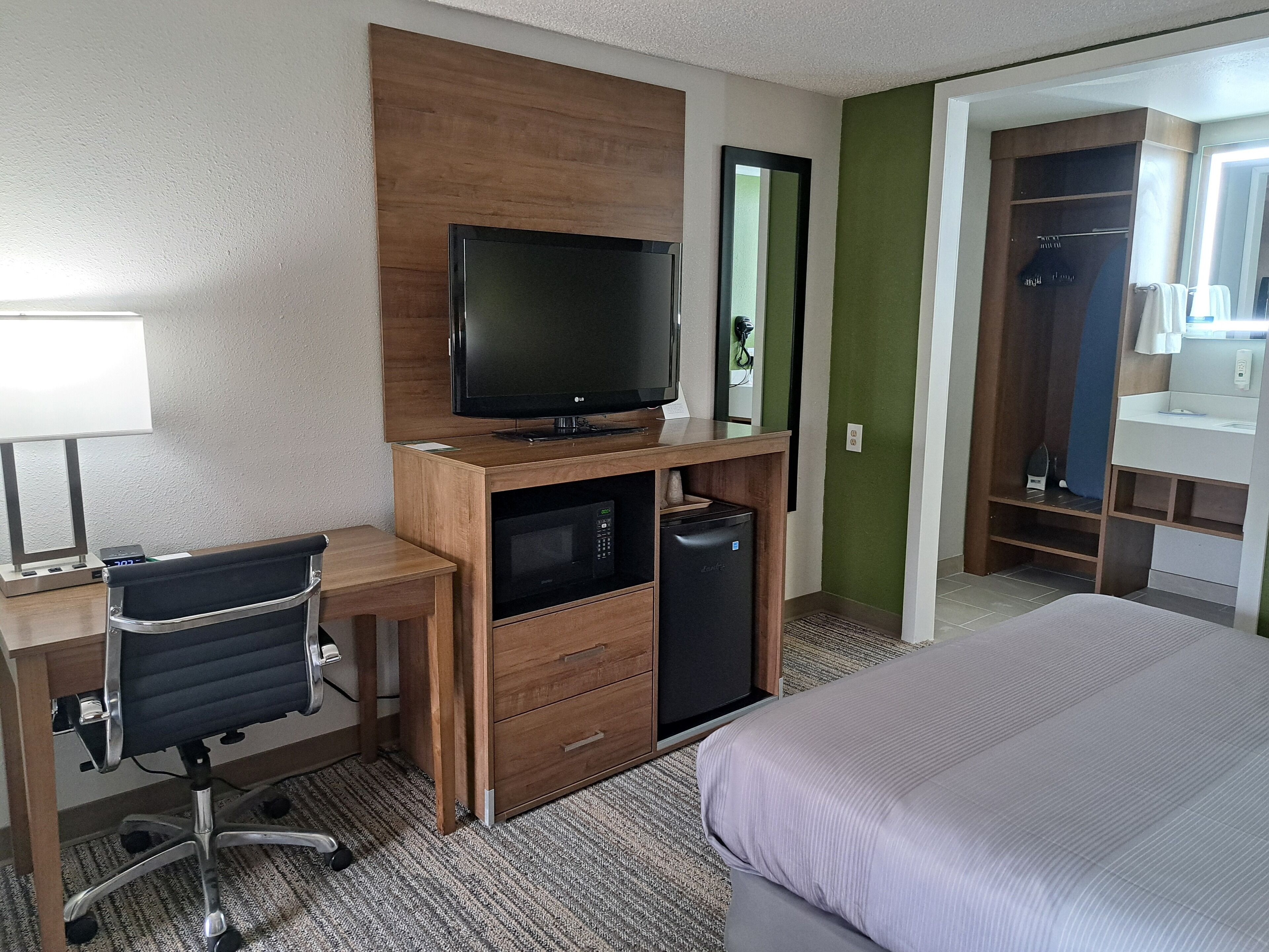 Hotelkamer met een houten bureau, een zwarte bureaustoel, een televisie, een magnetron, een minibar en een bed met een grijze plaid, plus een groene accentmuur.