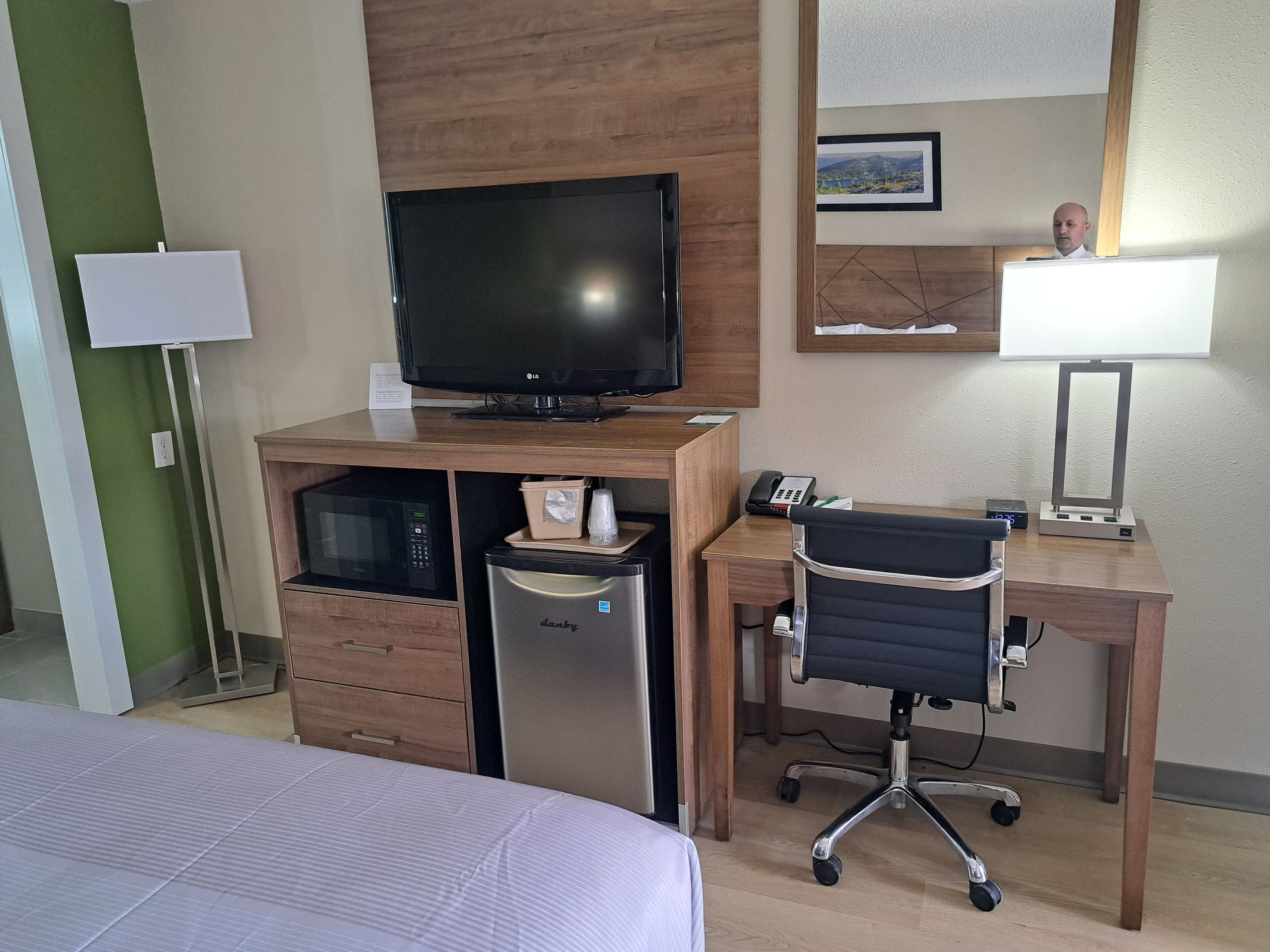 A hotel room with a bed, a desk with a computer, a television, a microwave, and a refrigerator.