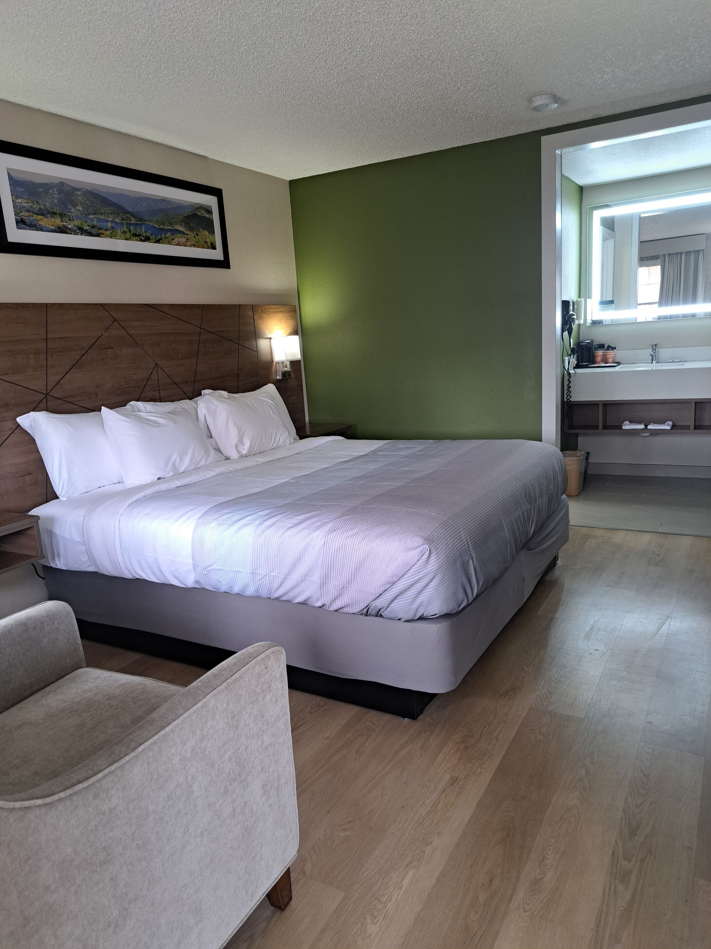 A modern hotel room with a large bed, a wooden headboard, a green accent wall, a bathroom with a mirror and sink, and a framed landscape picture on the wall.