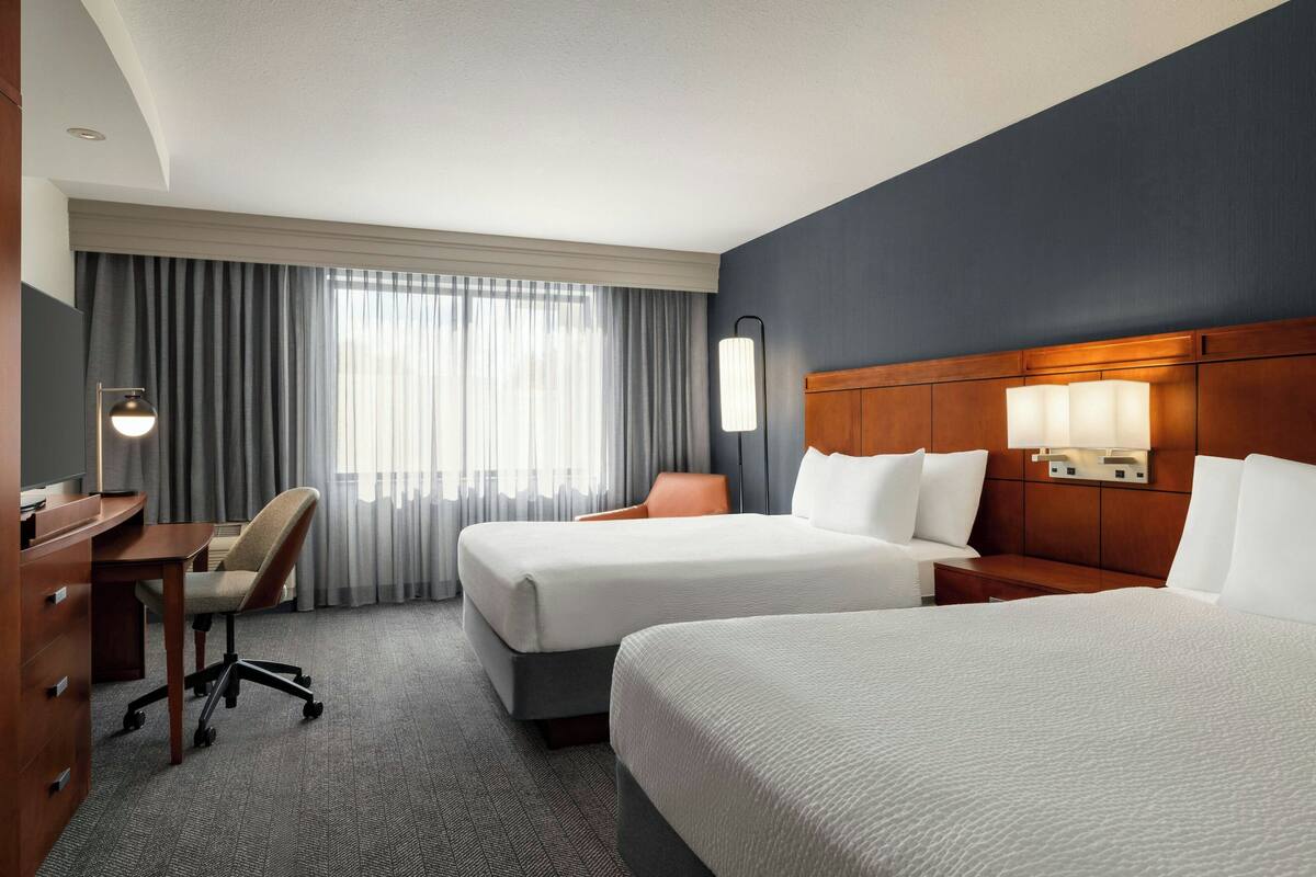 A hotel room with two beds, a desk, a chair, and a large window with curtains.