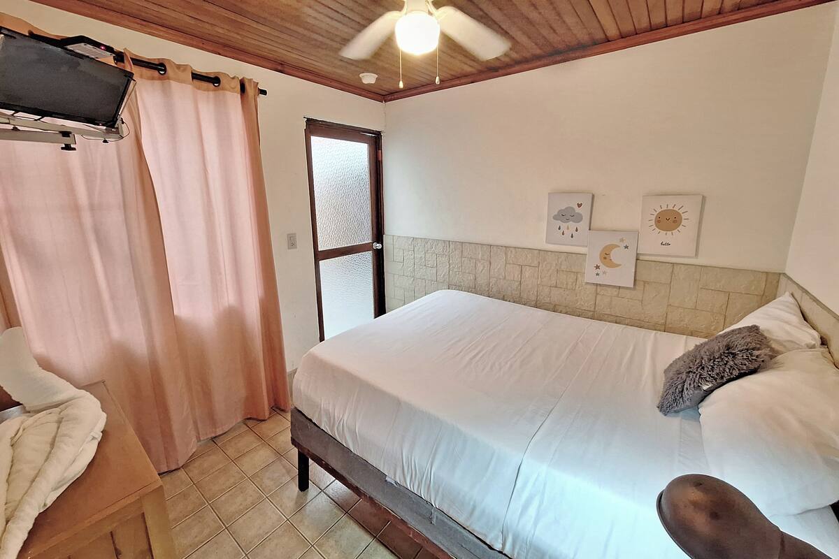 Een slaapkamer met een bed, een plafondventilator, een raam en een aan de muur bevestigde televisie.