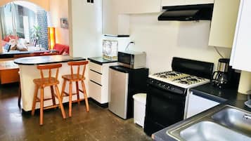 A compact kitchen with a gas stove, a microwave, a sink, and a small refrigerator. There are two wooden stools at a small table.
