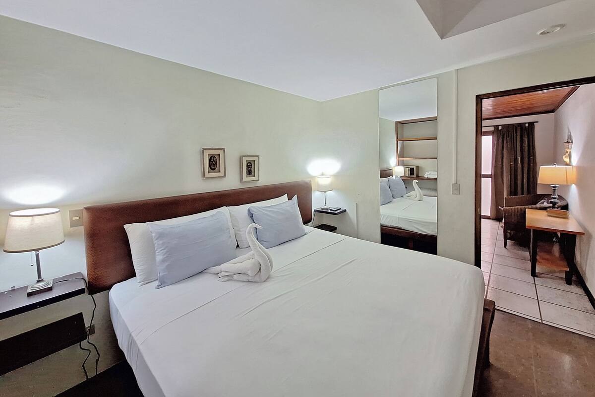 Een hotelkamer met een net opgemaakt bed, bedlampen, een spiegel en een klein tafeltje met een lamp.