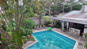 Piscina con sillas de descanso y zona de bar, rodeada de exuberante vegetación y árboles.