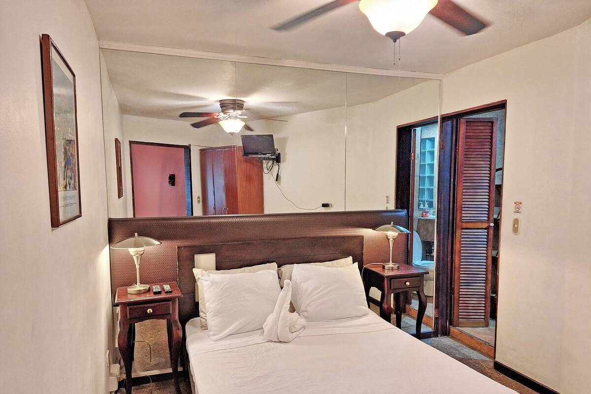 Een hotelkamer met een bed, nachtkastjes, een plafondventilator en een spiegel die de kamer weerspiegelt.
