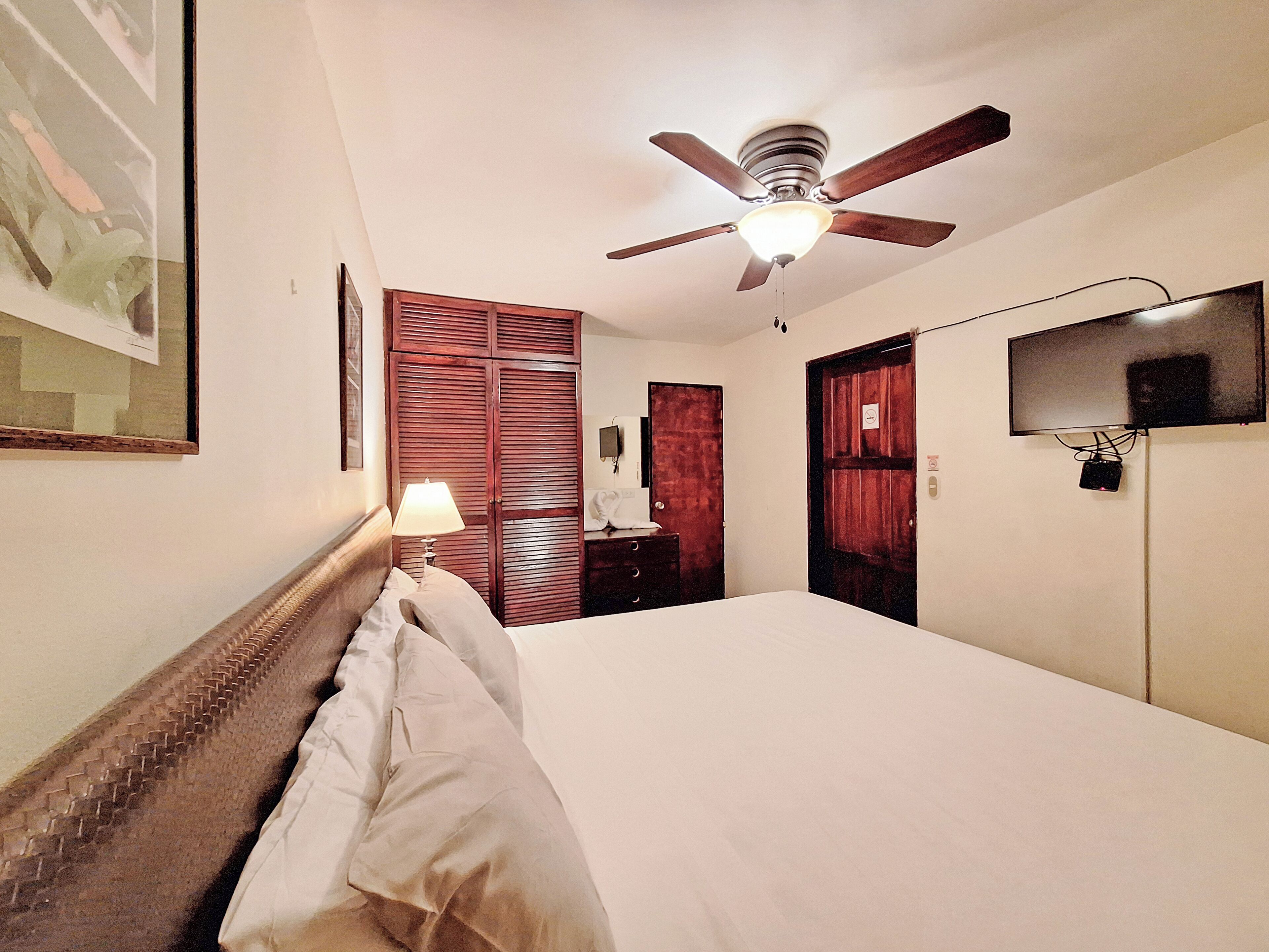 A hotel room with a bed, a ceiling fan, a TV mounted on the wall, and a wooden wardrobe.