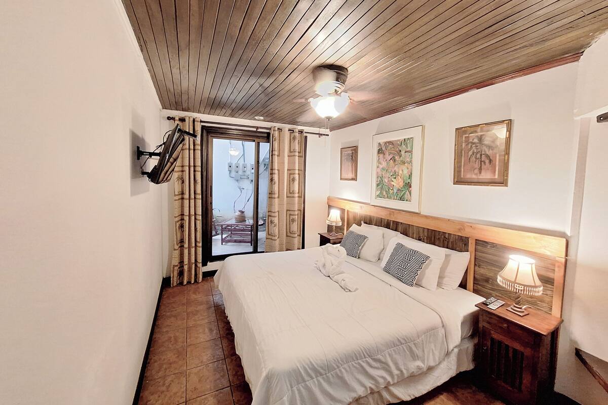 Een slaapkamer met een houten plafond, een bed met wit beddengoed, een nachtkastje met een lamp en een raam met gordijnen.
