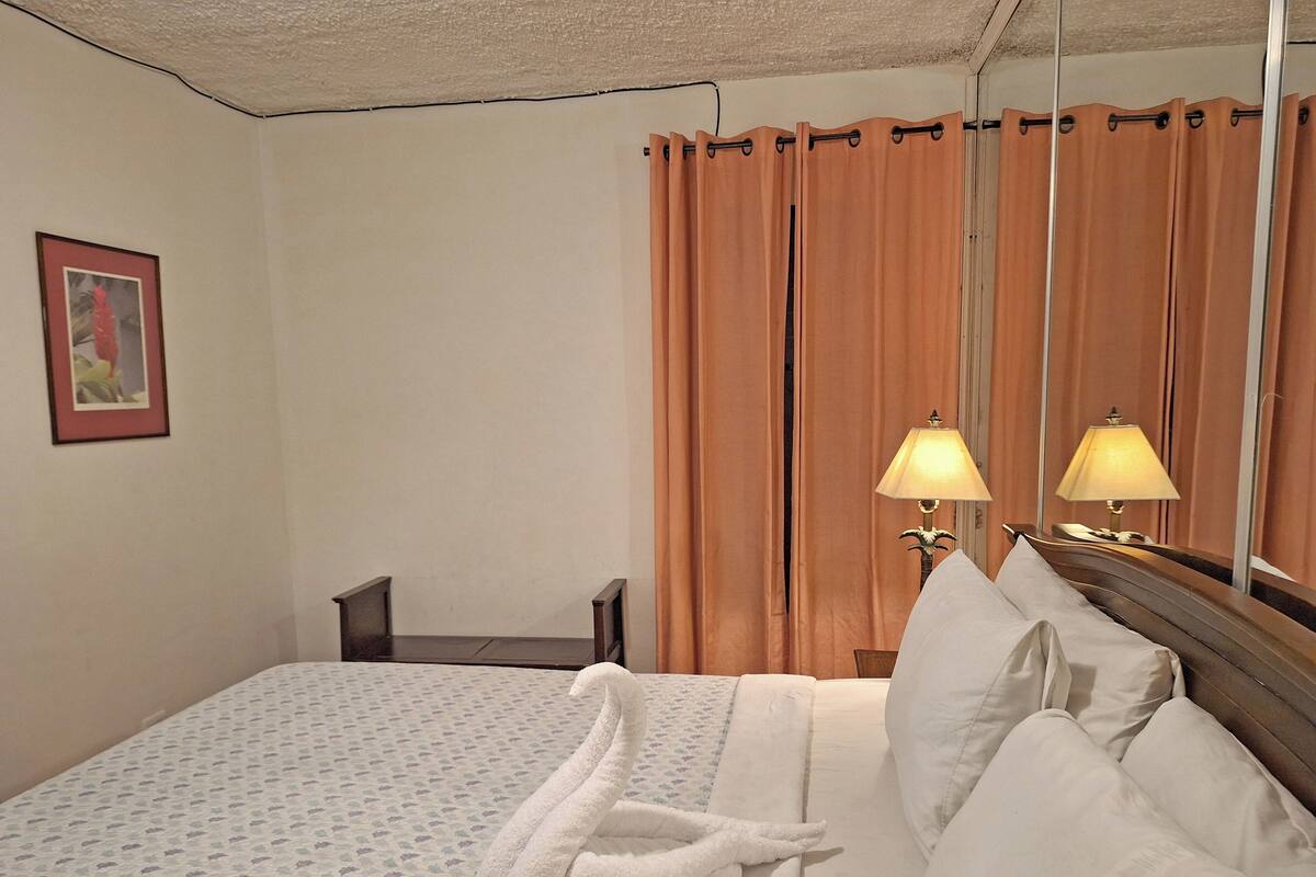 Een hotelkamer met een bed, twee bedlampen, een spiegel, een inlijsting foto aan de muur en een raam met gordijnen.
