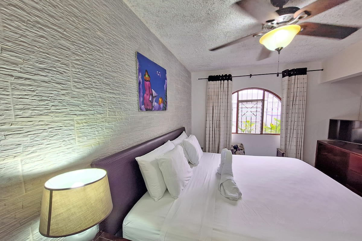 Een hotelkamer met een bed, een plafondventilator, een raam met gordijnen en een schilderij aan de muur.