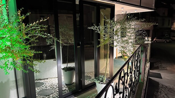Une vue nocturne d’un bâtiment doté de grandes fenêtres, de plantes en pot et d’une rambarde en métal.