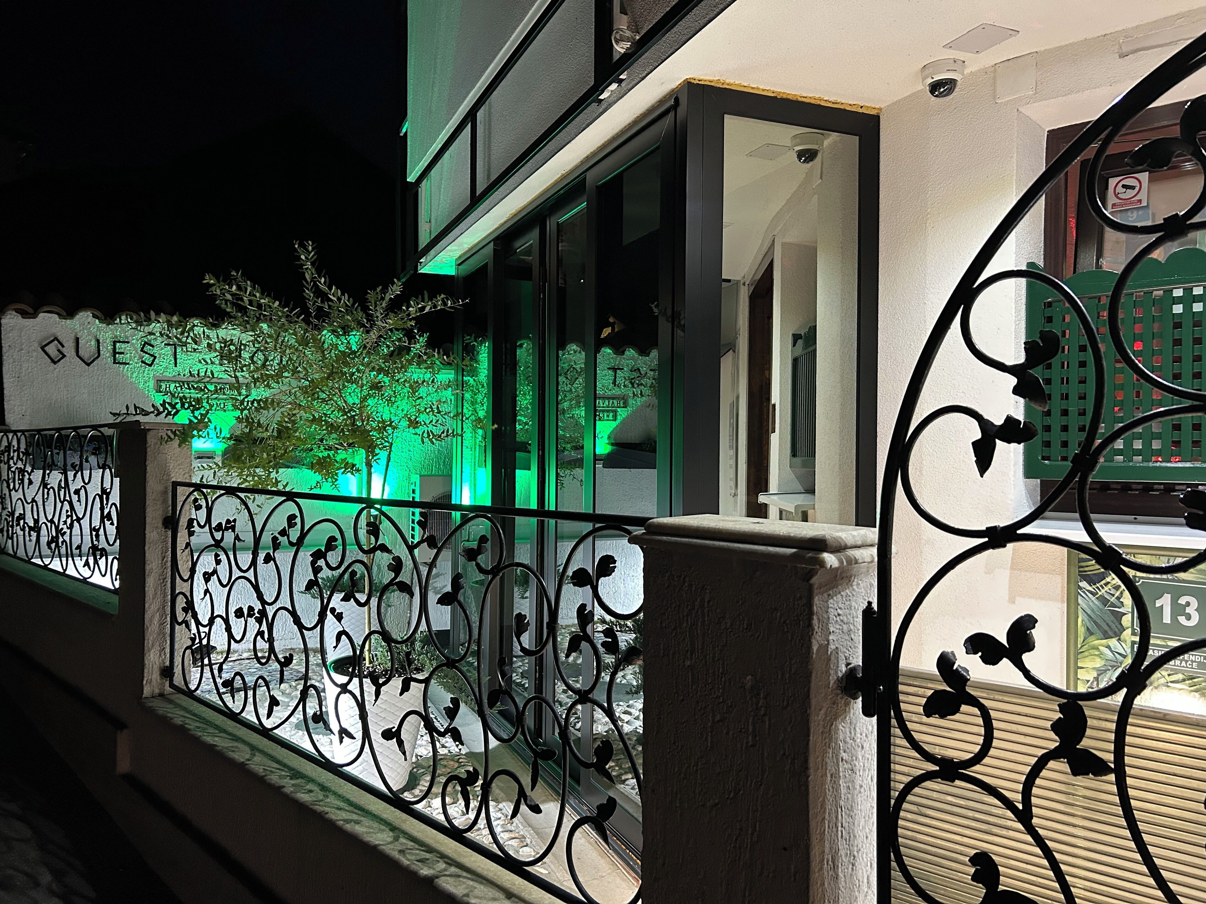 A hotel entrance with a decorative iron gate, a visible sign reading "GUESTS," and a green-lit exterior.