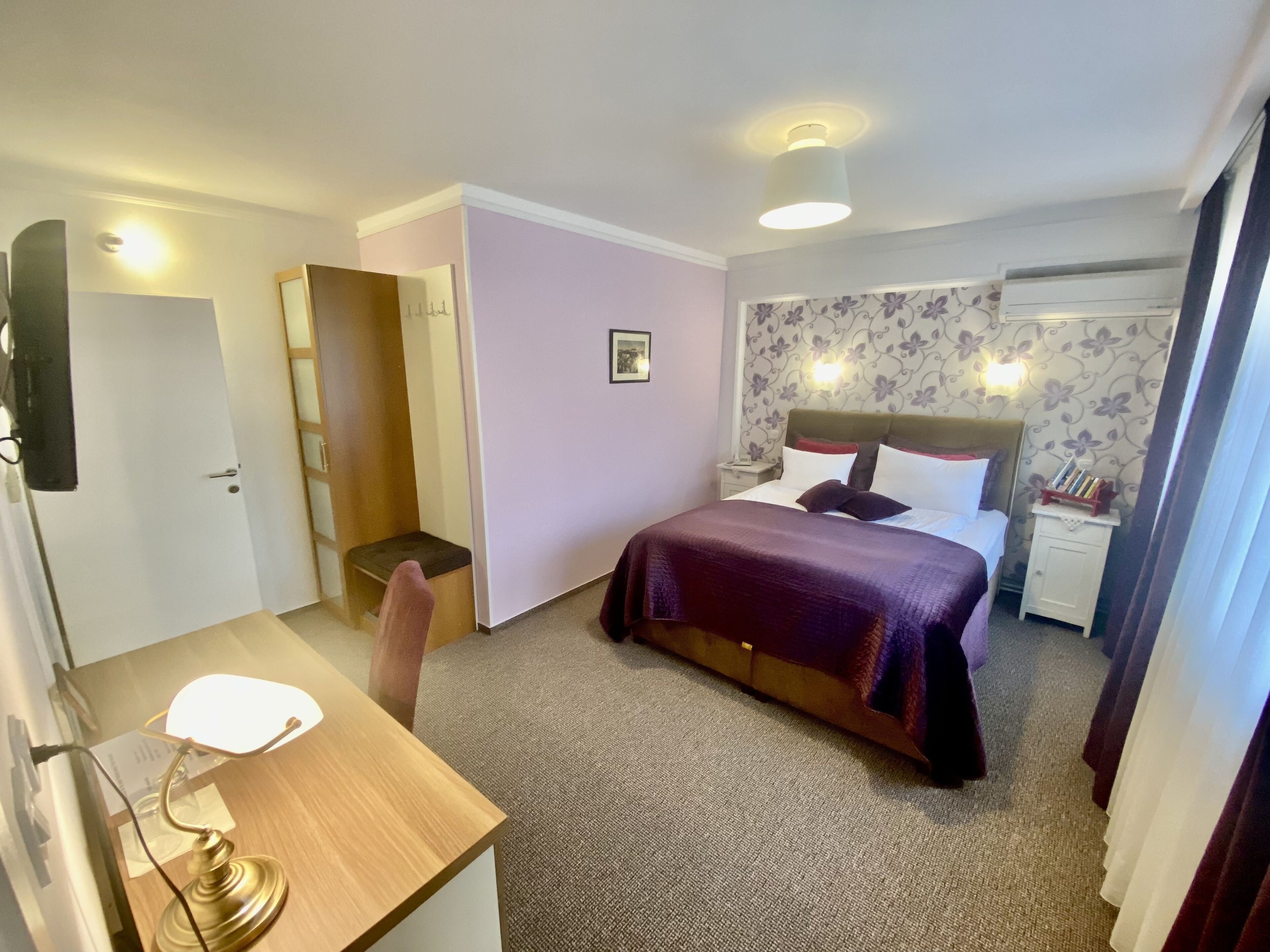 Quarto de hotel com cama, mesas de cabeceira, escrivaninha com abajur, cadeira, guarda-roupa e porta.