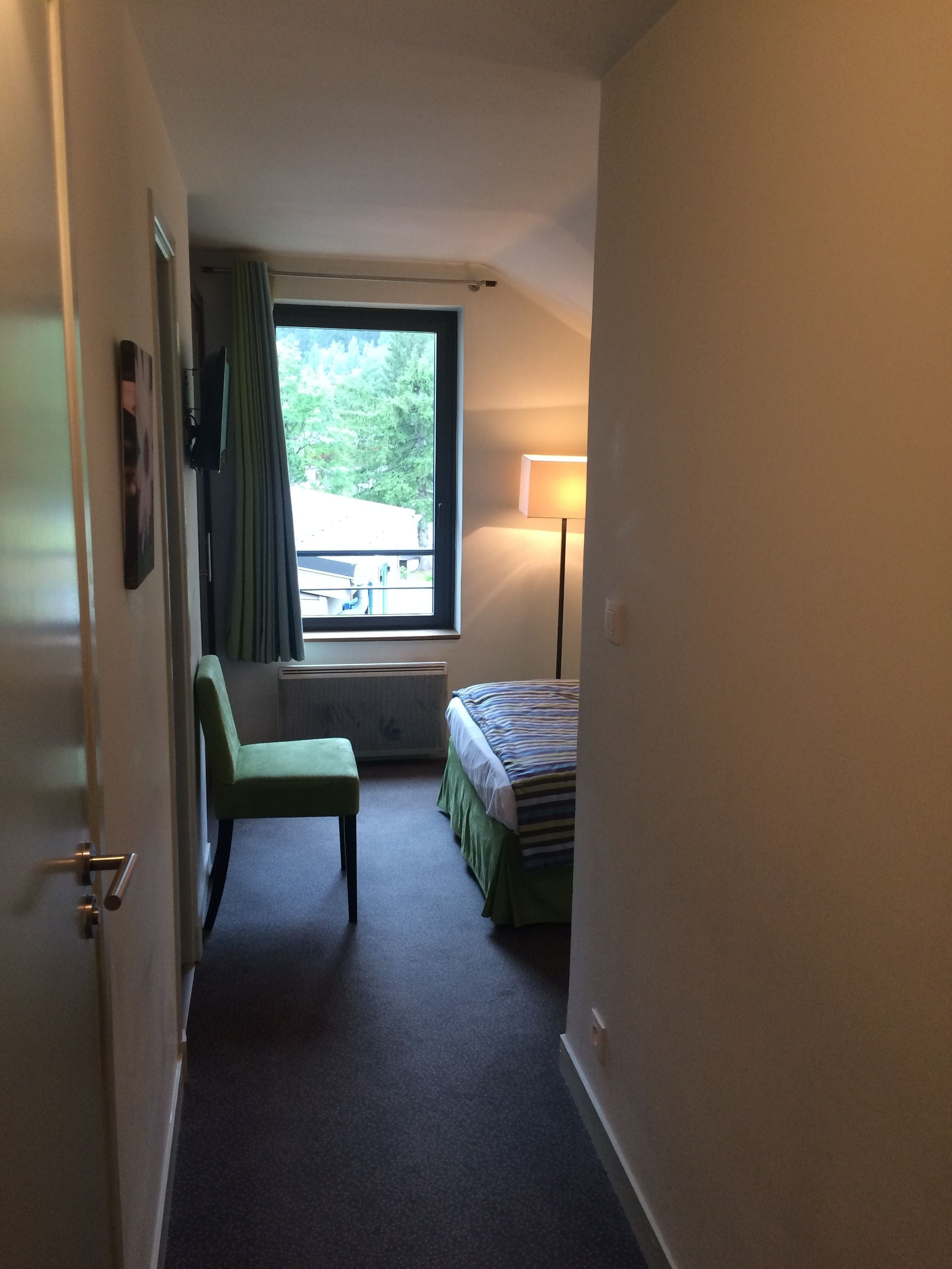 Habitación de hotel estrecha con cama, un escritorio pequeño, una silla, ventana con cortinas y una lámpara.