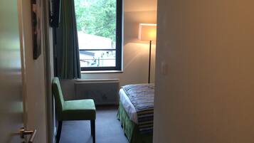 Habitación de hotel estrecha con cama, un escritorio pequeño, una silla, ventana con cortinas y una lámpara.