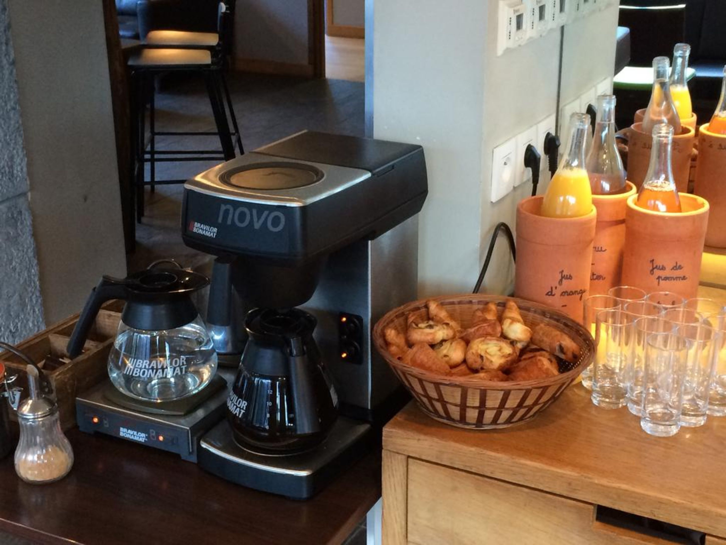 A coffee station with a coffee machine, a basket of pastries, and a row of glasses and cups.