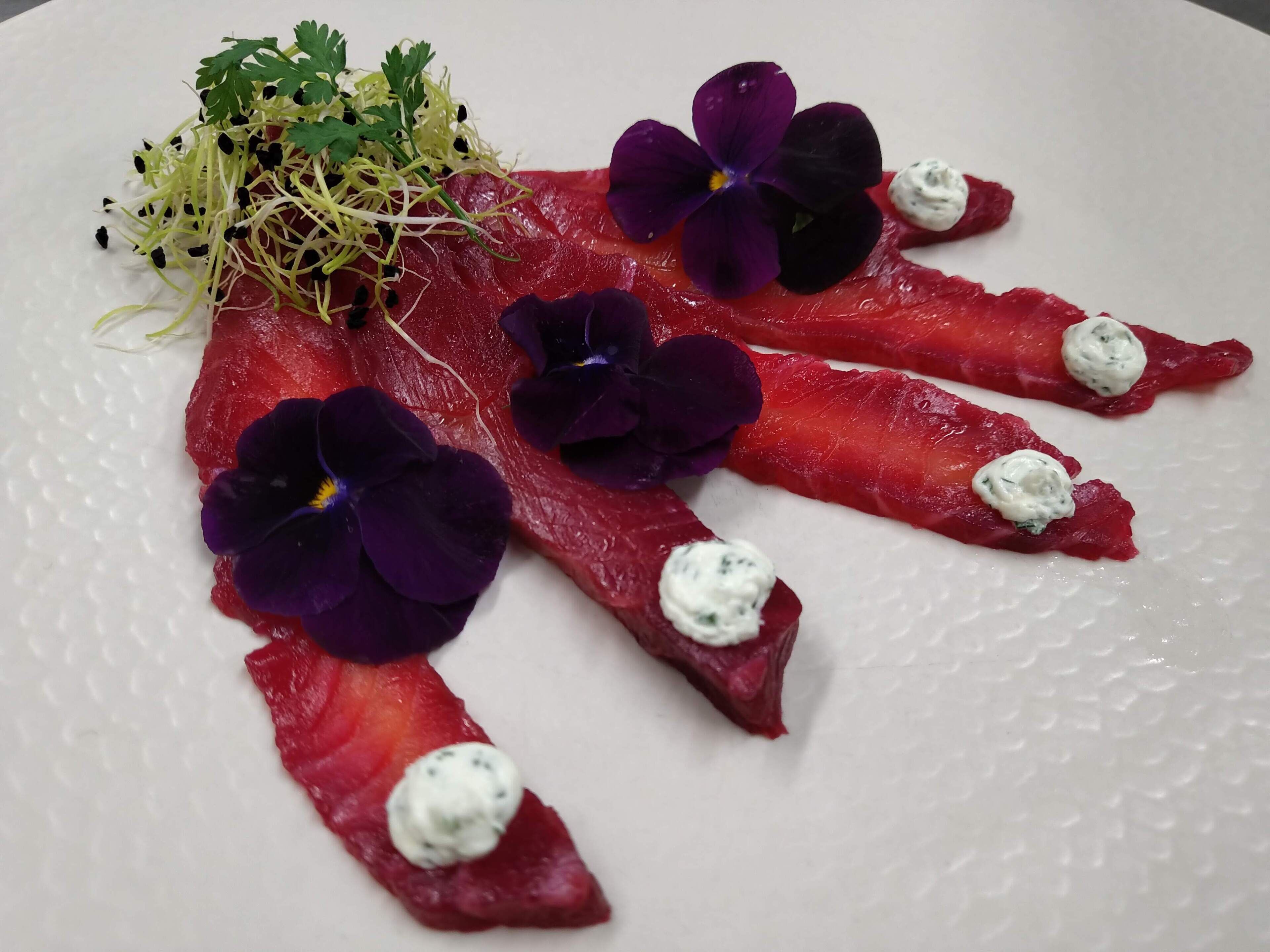 Un plato con carne roja cortada en lonjas finas, adornado con flores púrpuras y un pequeño adorno verde.