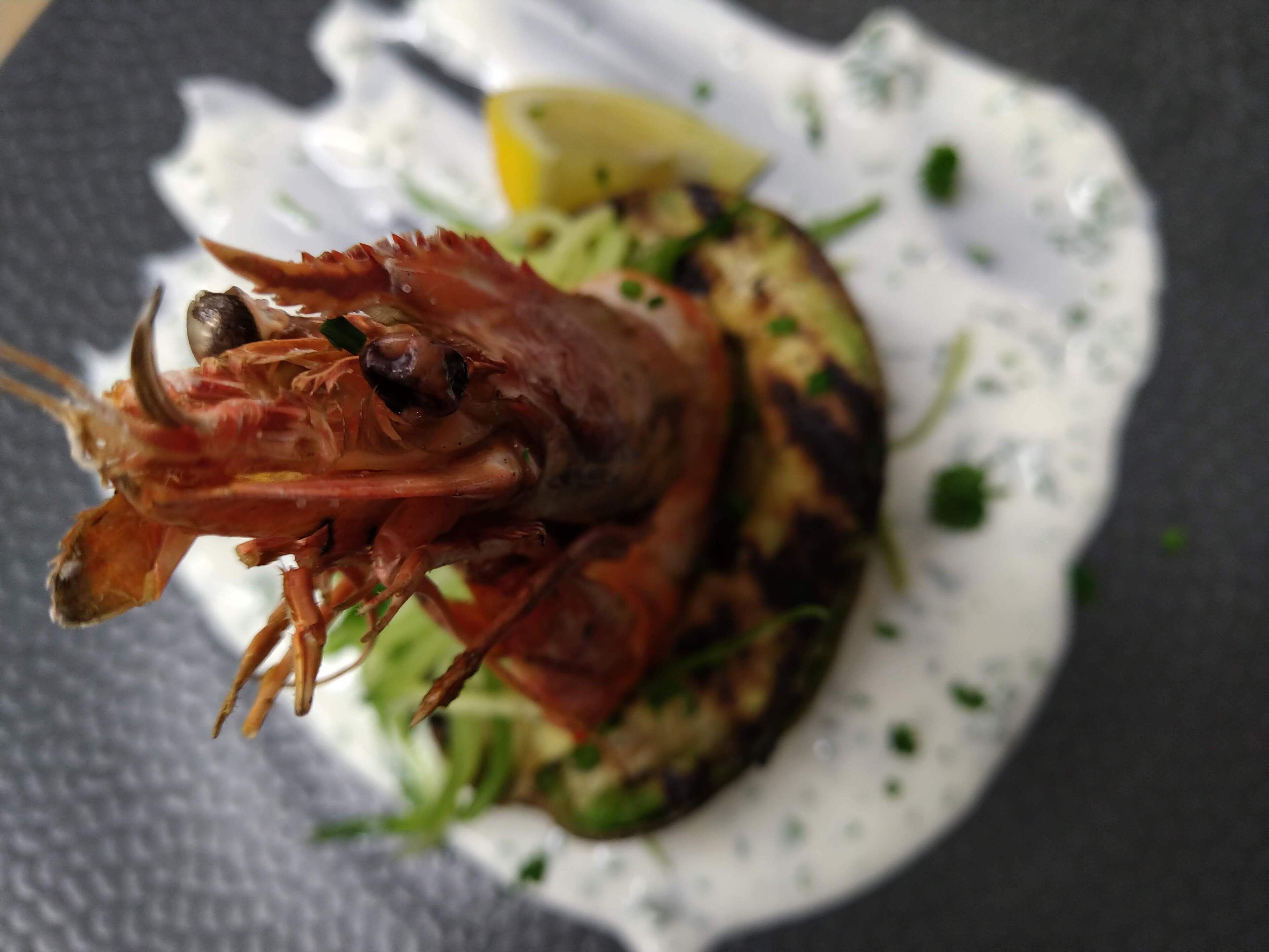 Un langostino a la parrilla sobre una cama de vegetales verdes, con un gajo de limón y salsa cremosa.
