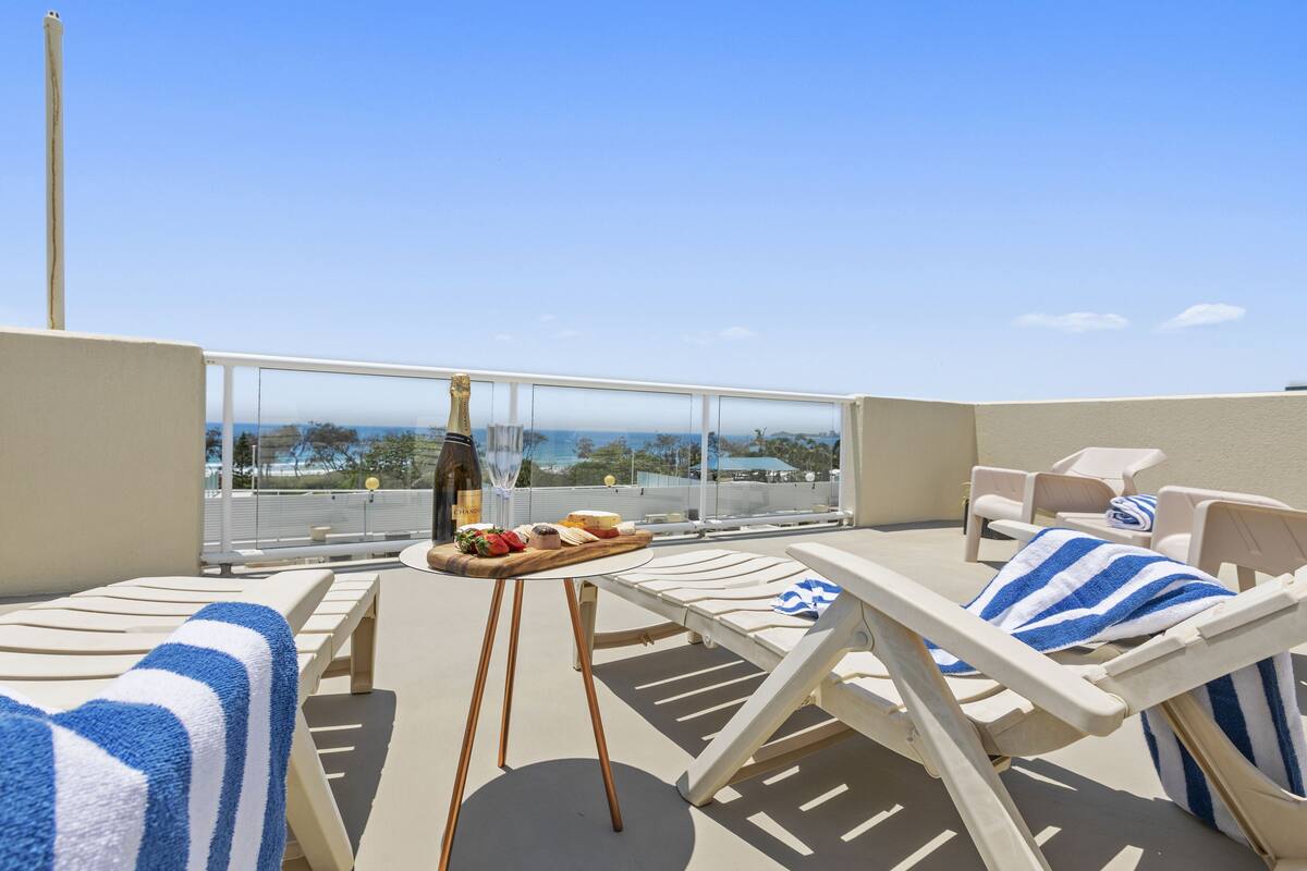A rooftop terrace with lounge chairs, a table with food and drinks, and a view of the ocean.