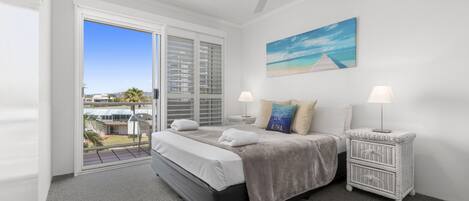 A bedroom with a bed, two bedside tables with lamps, a painting of a beach scene, and a view of a house and palm trees through the window.