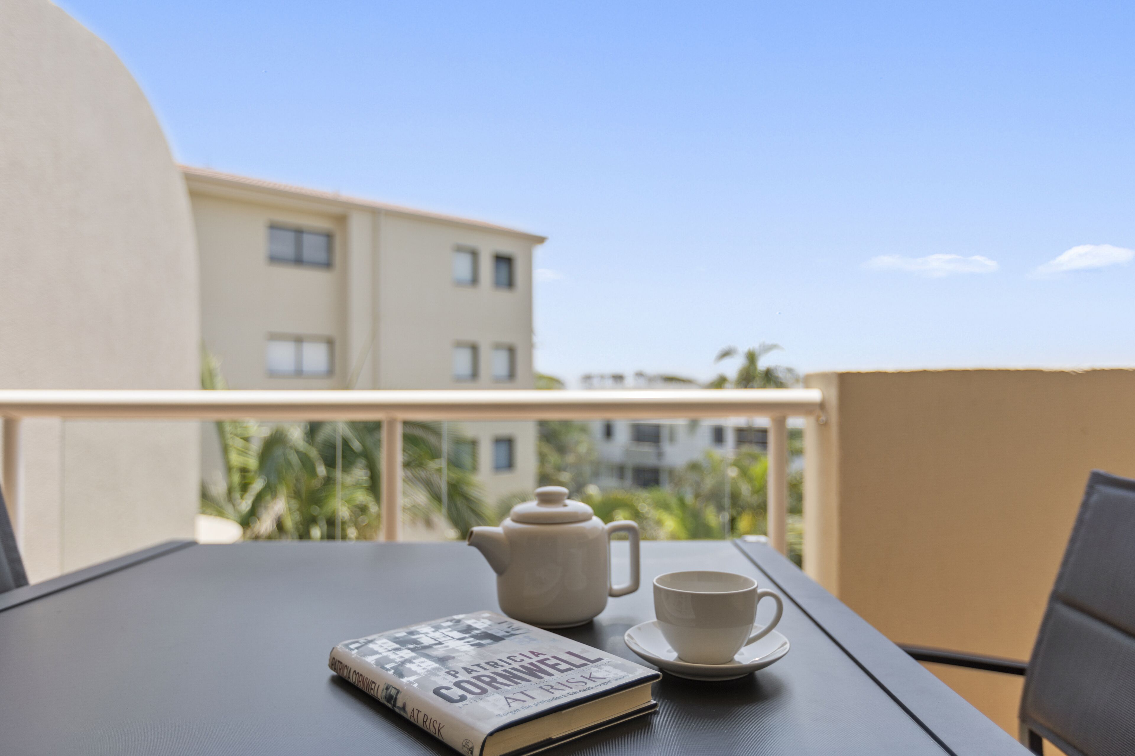 Een tafel met een boek, theepot en kopje op een balkon met uitzicht op een gebouw en groen.