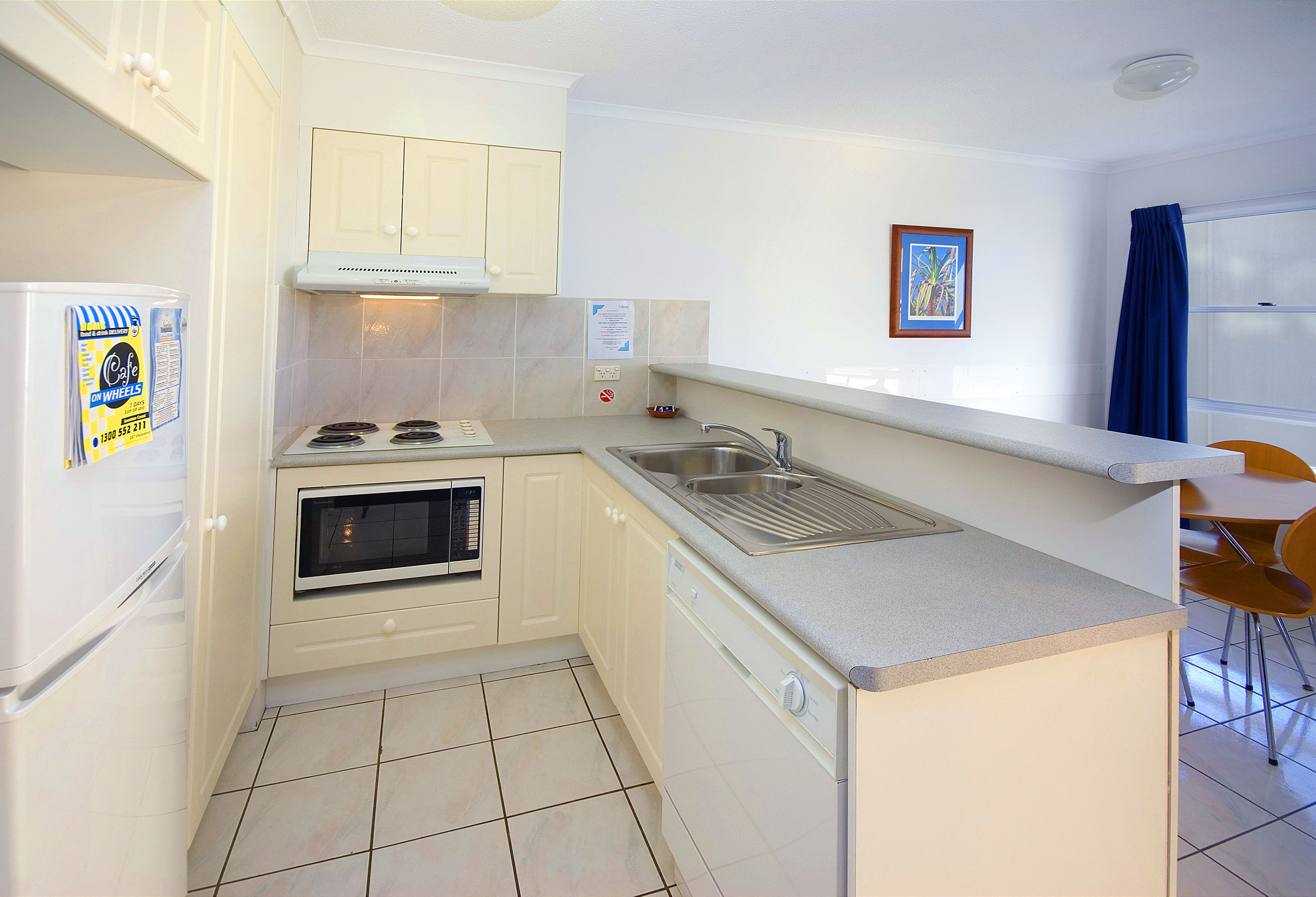 A compact kitchen with white cabinets, a built-in oven, a dishwasher, and a sink. There is a window with blue curtains and a framed picture on the wall.