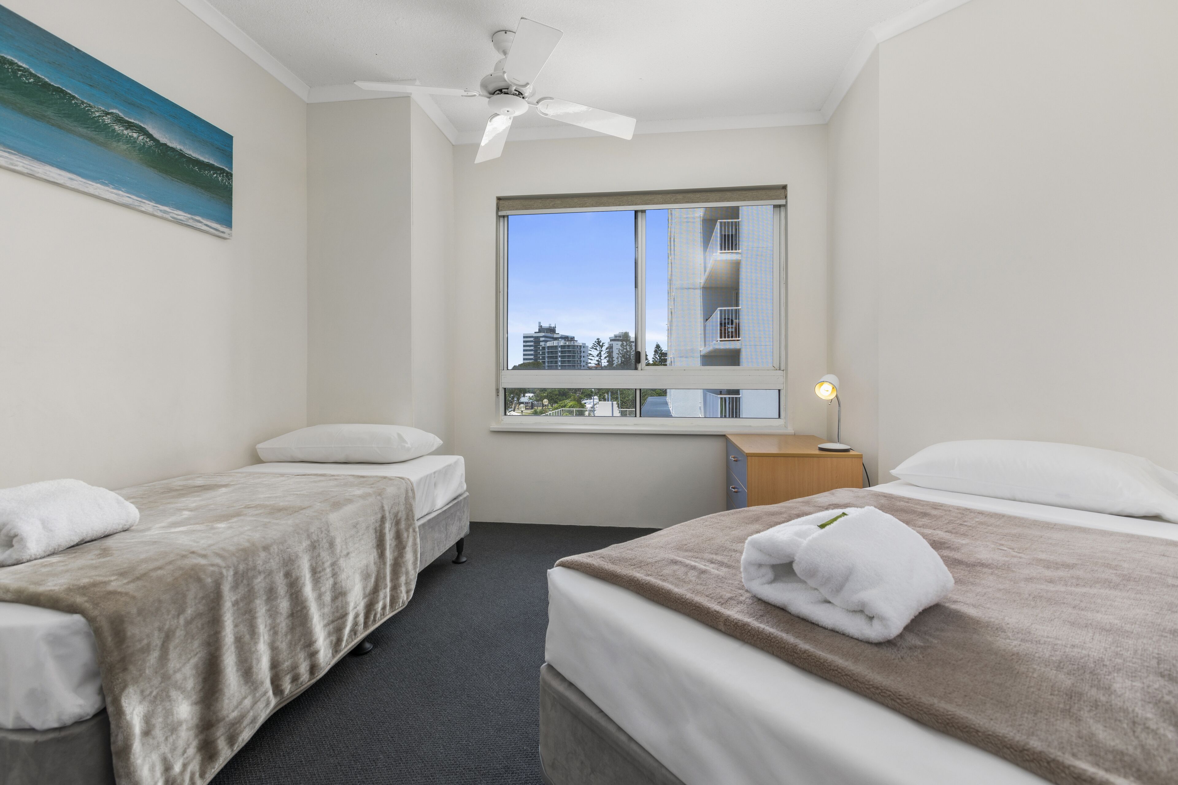 Una camera d'albergo con due letti, una ventola a soffitto, una finestra con vista sulla città e un quadro appeso al muro.