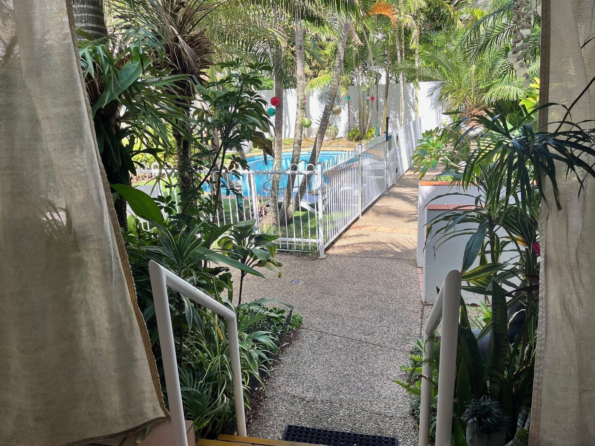 A view of a pool area with a white fence, surrounded by various plants and trees.