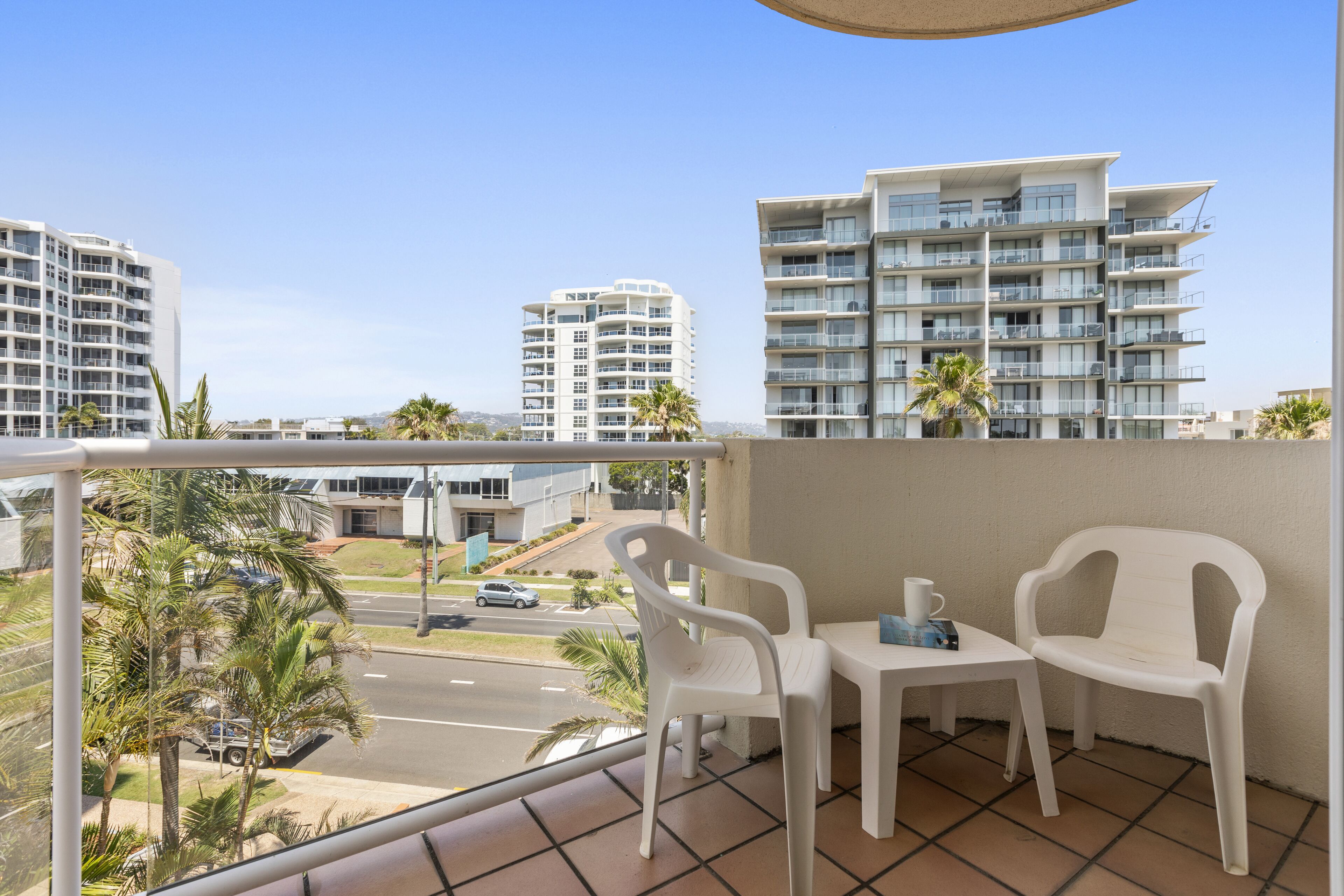 A balcony with two white plastic chairs and a small table, overlooking a street with palm trees and modern buildings.