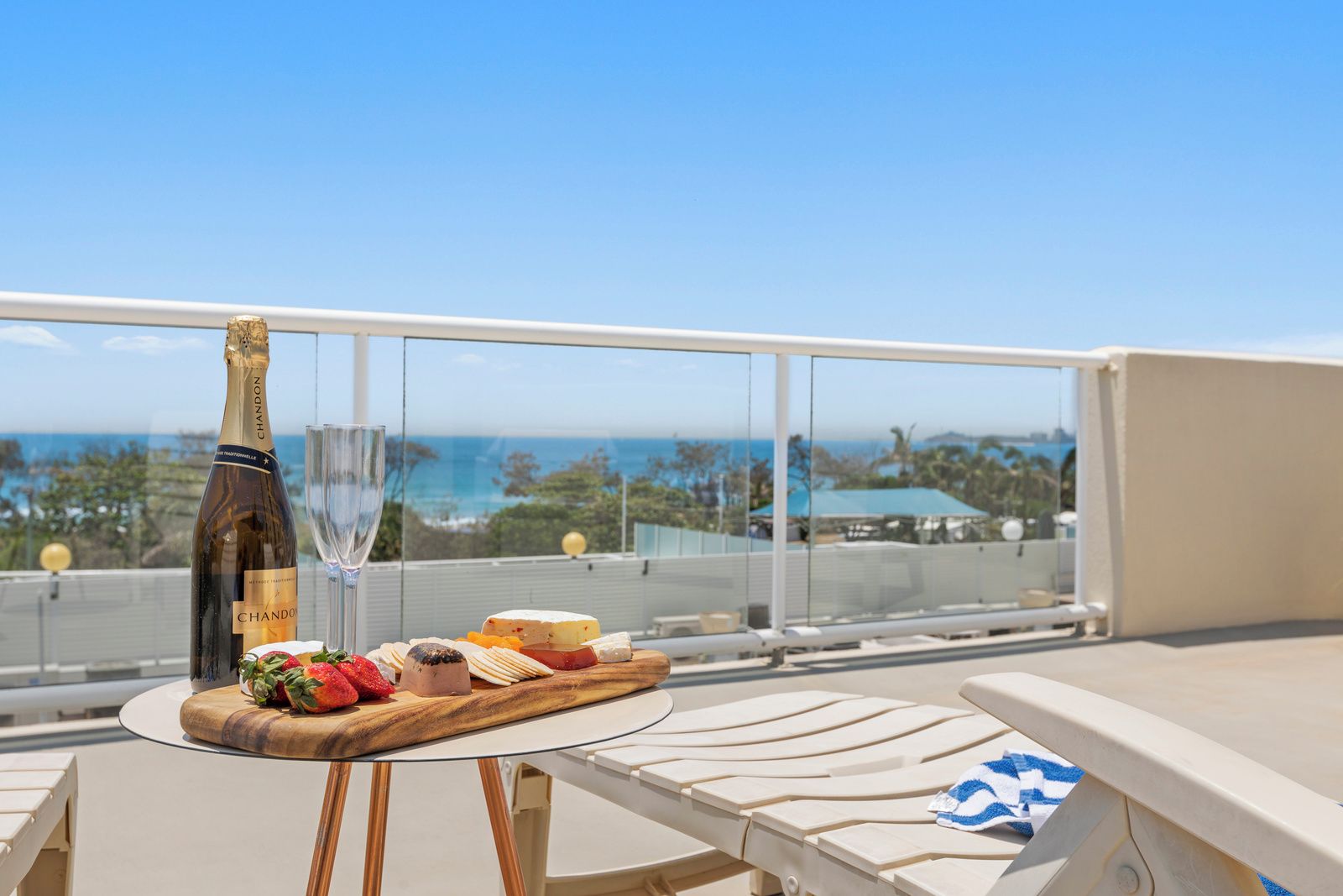 A balcony with a table set for a meal, a bottle of wine, and a glass. The balcony overlooks a scenic view of the ocean and a distant coastline.