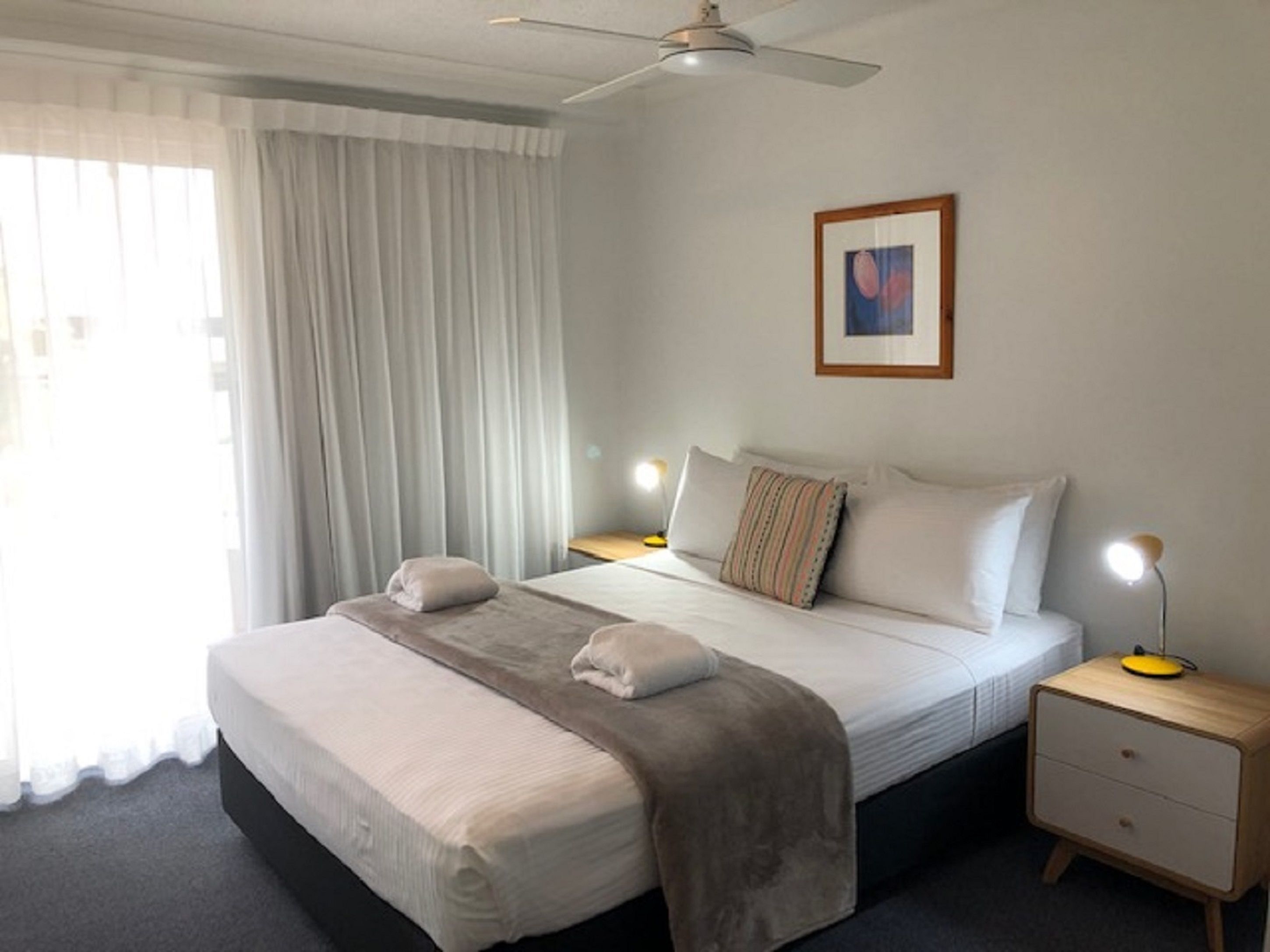 A neatly made bed with a grey blanket, two bedside lamps, a framed picture on the wall, and a ceiling fan.