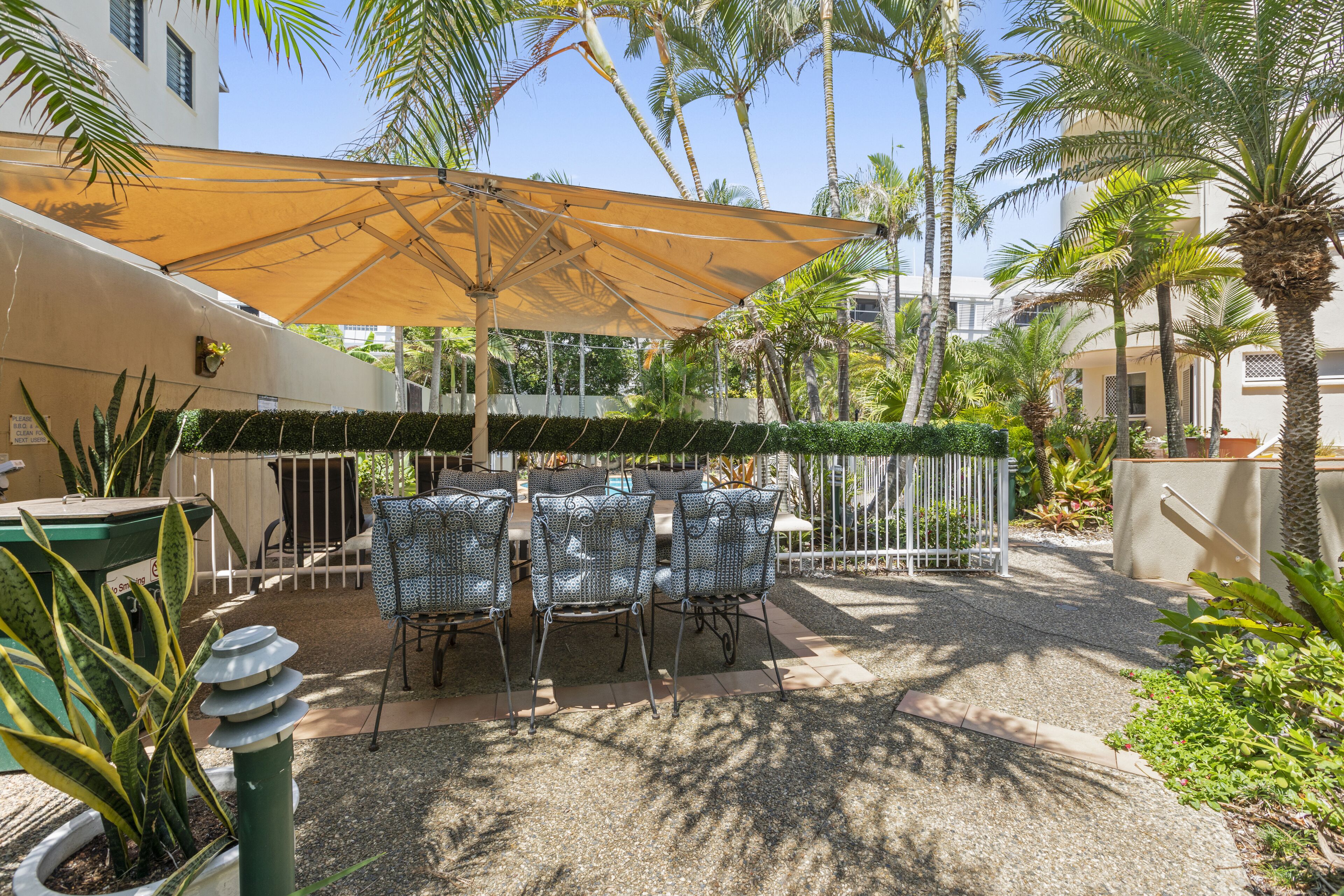 Eine Außenpatio-Terrasse mit Tisch und Stühlen unter einem großen Sonnenschirm, umgeben von Palmen und Grünpflanzen.