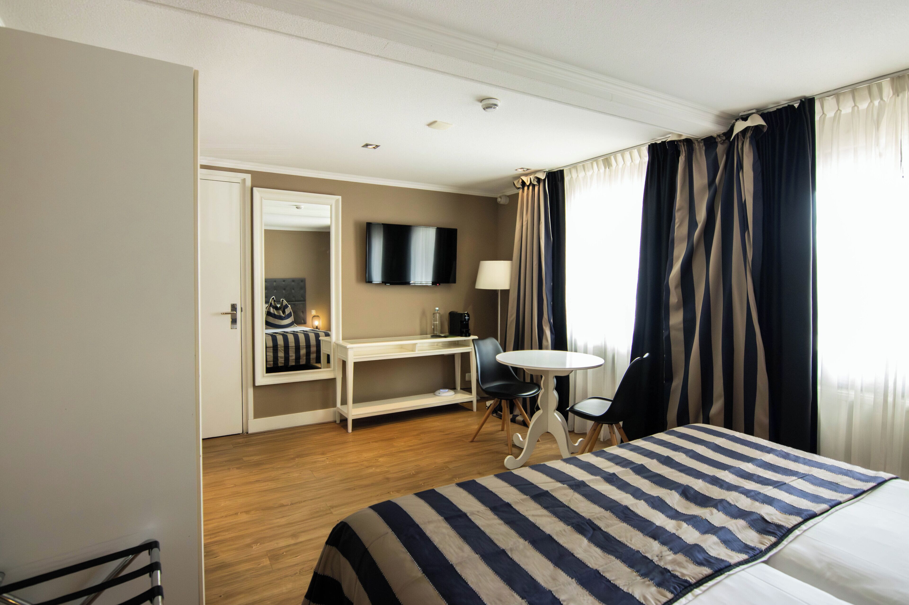 Una habitación de hotel con cama, escritorio, silla, televisión y ventana con cortinas.