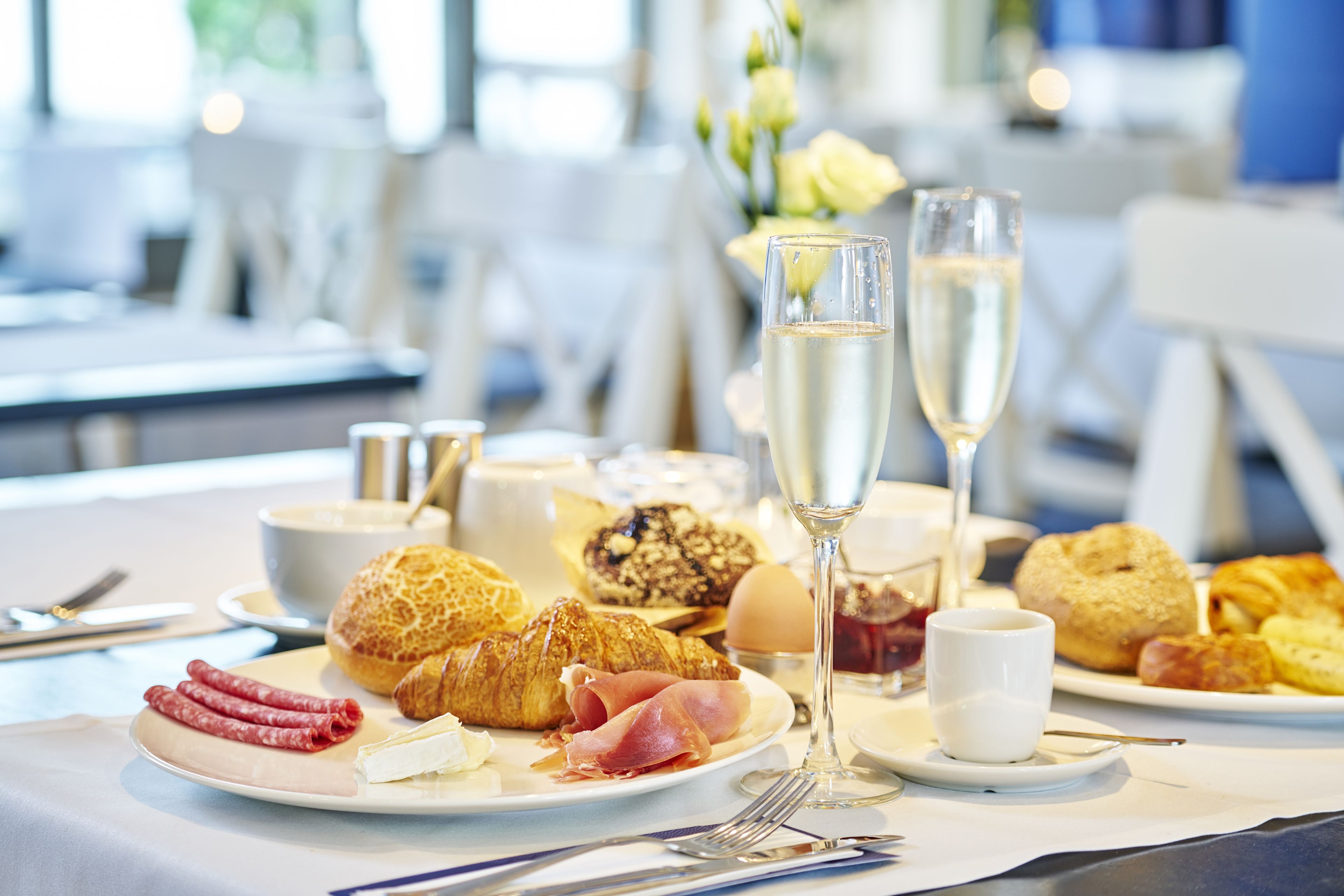 Una escena de desayuno con croissants, jamón, huevos y copas de champán sobre una mesa.