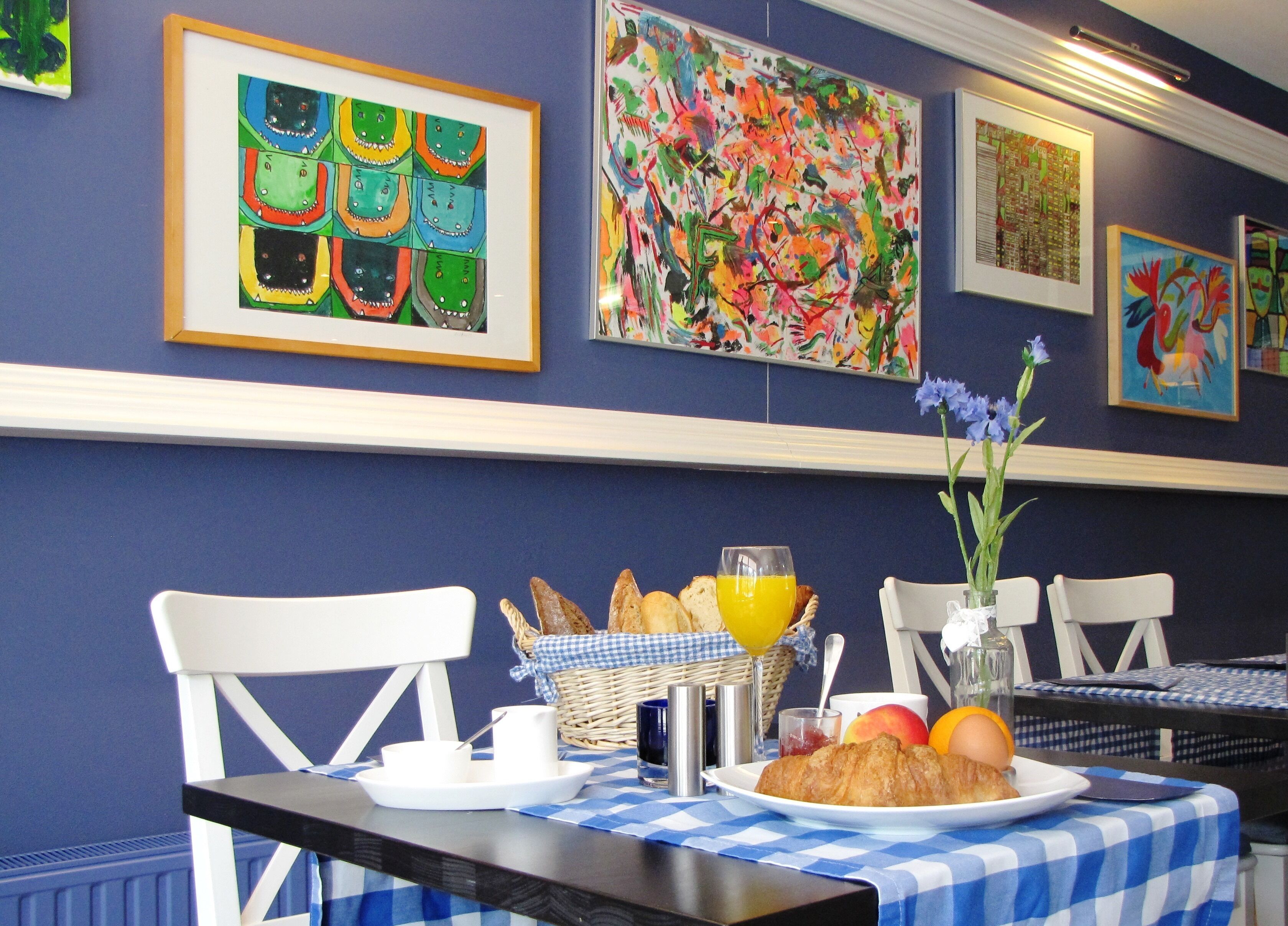 Une table de petit-déjeuner avec un croissant, du jus d’orange et des fruits, placée devant un mur orné d’œuvres d’art colorées.