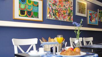 Une table de petit-déjeuner avec un croissant, du jus d’orange et des fruits, placée devant un mur orné d’œuvres d’art colorées.