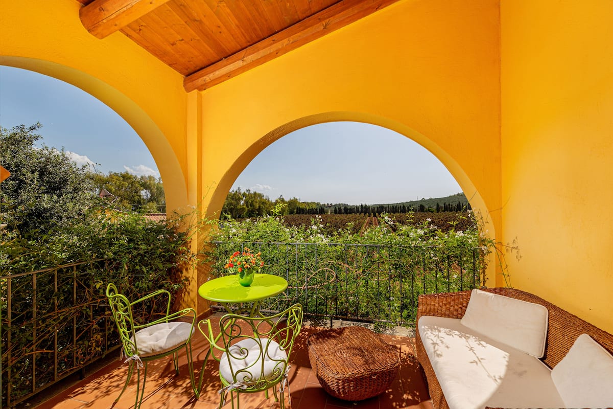 Un balcon avec un mur jaune, un canapé en osier, une chaise en métal verte et une petite table sur laquelle est posé un vase de fleurs.