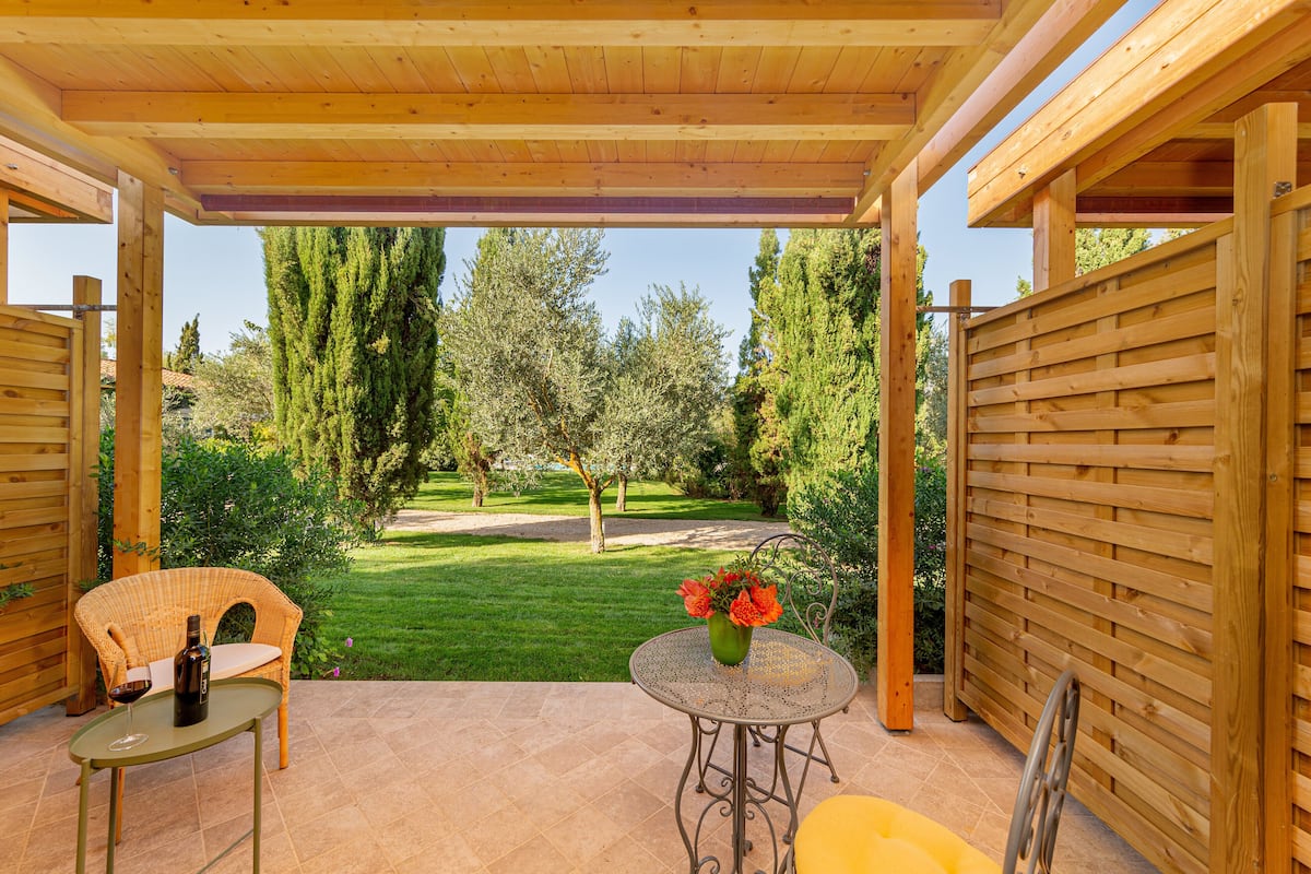 Une terrasse extérieure agrémentée d’une pergola en bois, d’un fauteuil en osier, d’une petite table ronde ornée d’un vase de fleurs et d’une bouteille de vin.