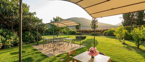 An outdoor patio with a table and chairs under a canopy, surrounded by well-maintained grass and bushes.