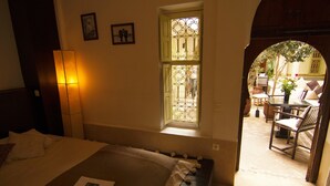 Une chambre d’hôtel avec un lit, une fenêtre ornée de barreaux en fer forgé, une petite table avec un vase, et une vue sur la cour.