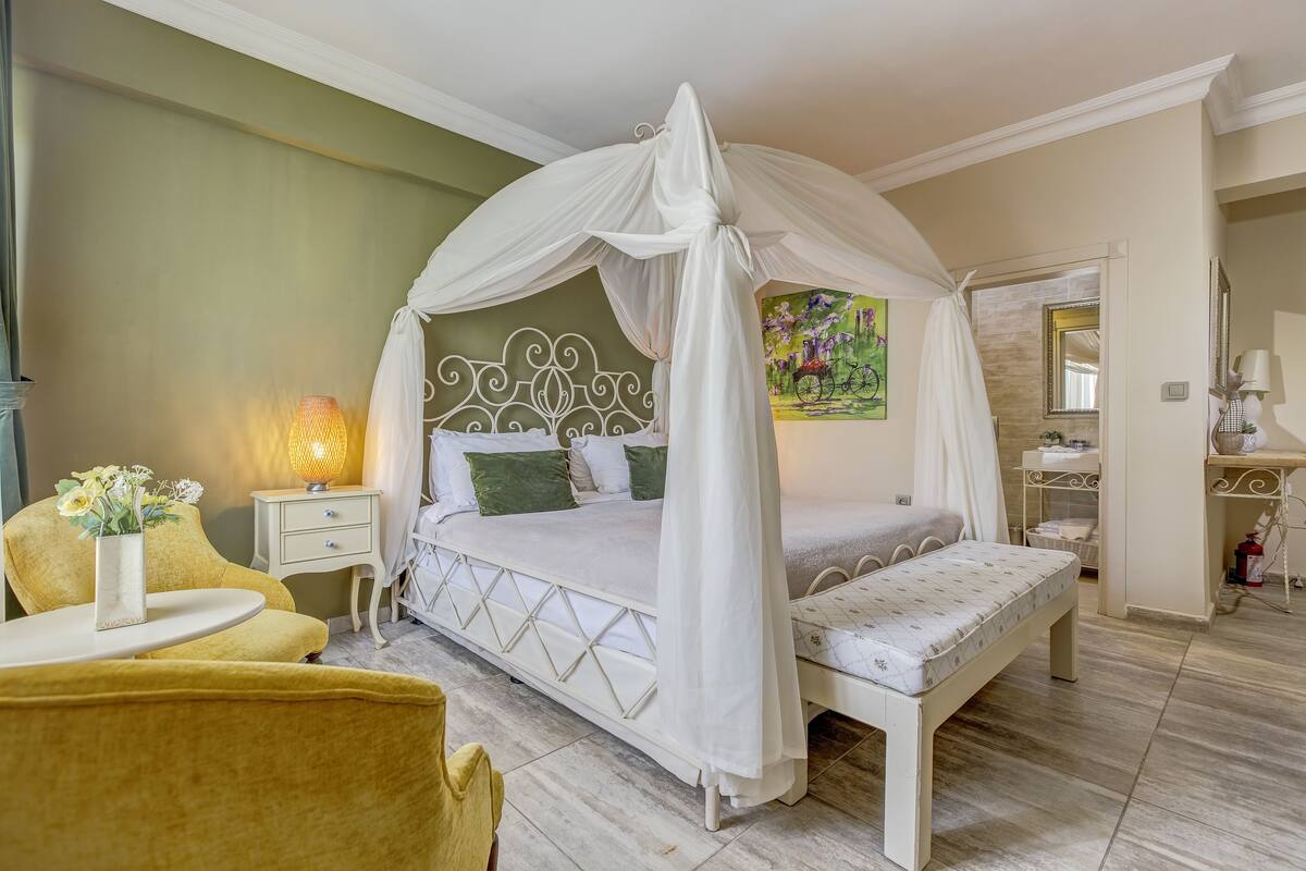 Un dormitorio con cama con dosel, una mesita de noche con lámpara, una silla y una pequeña mesa con un jarrón de flores.