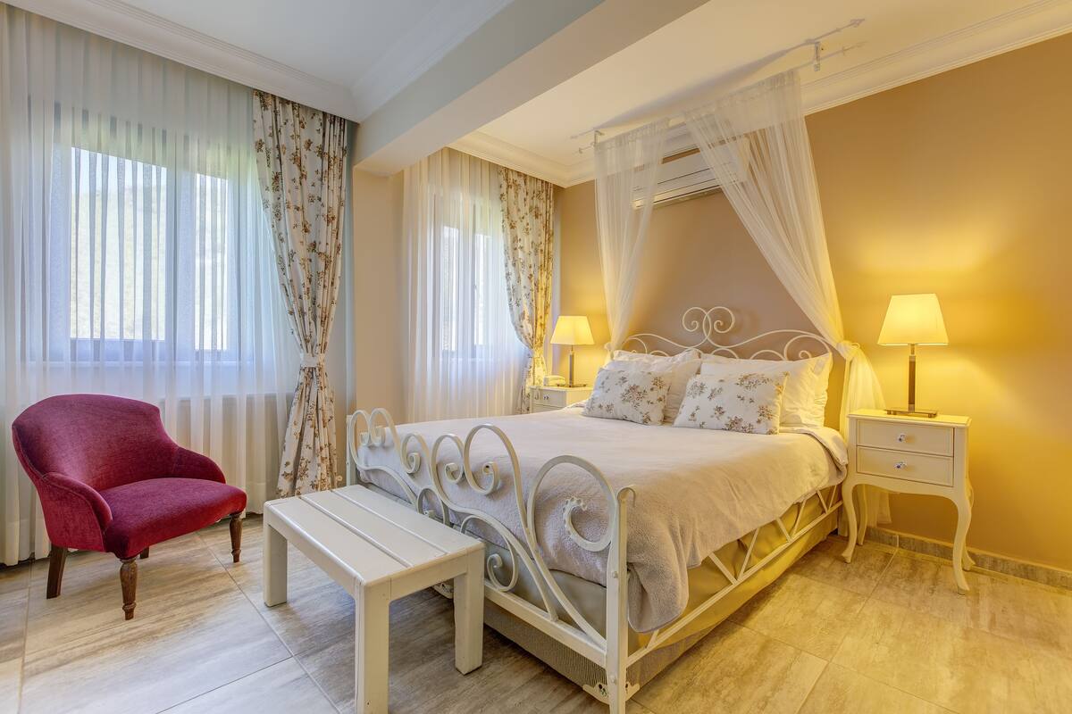 Un dormitorio con cama con dosel, una mesita de noche con lámpara, un sillón rojo y un banco.